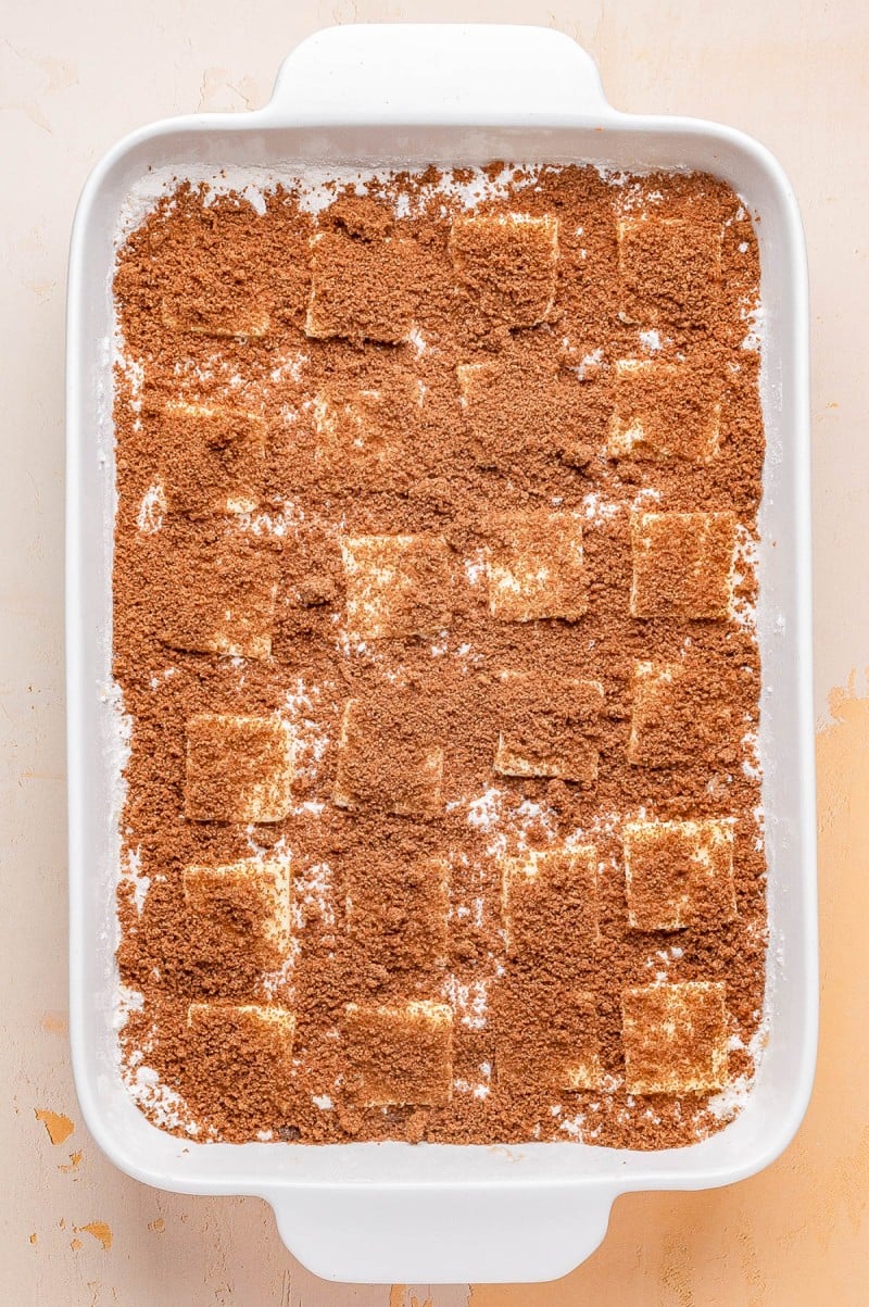 A white baking dish filled with layered rectangular pieces dusted with a generous amount of brown cinnamon sugar mixture.