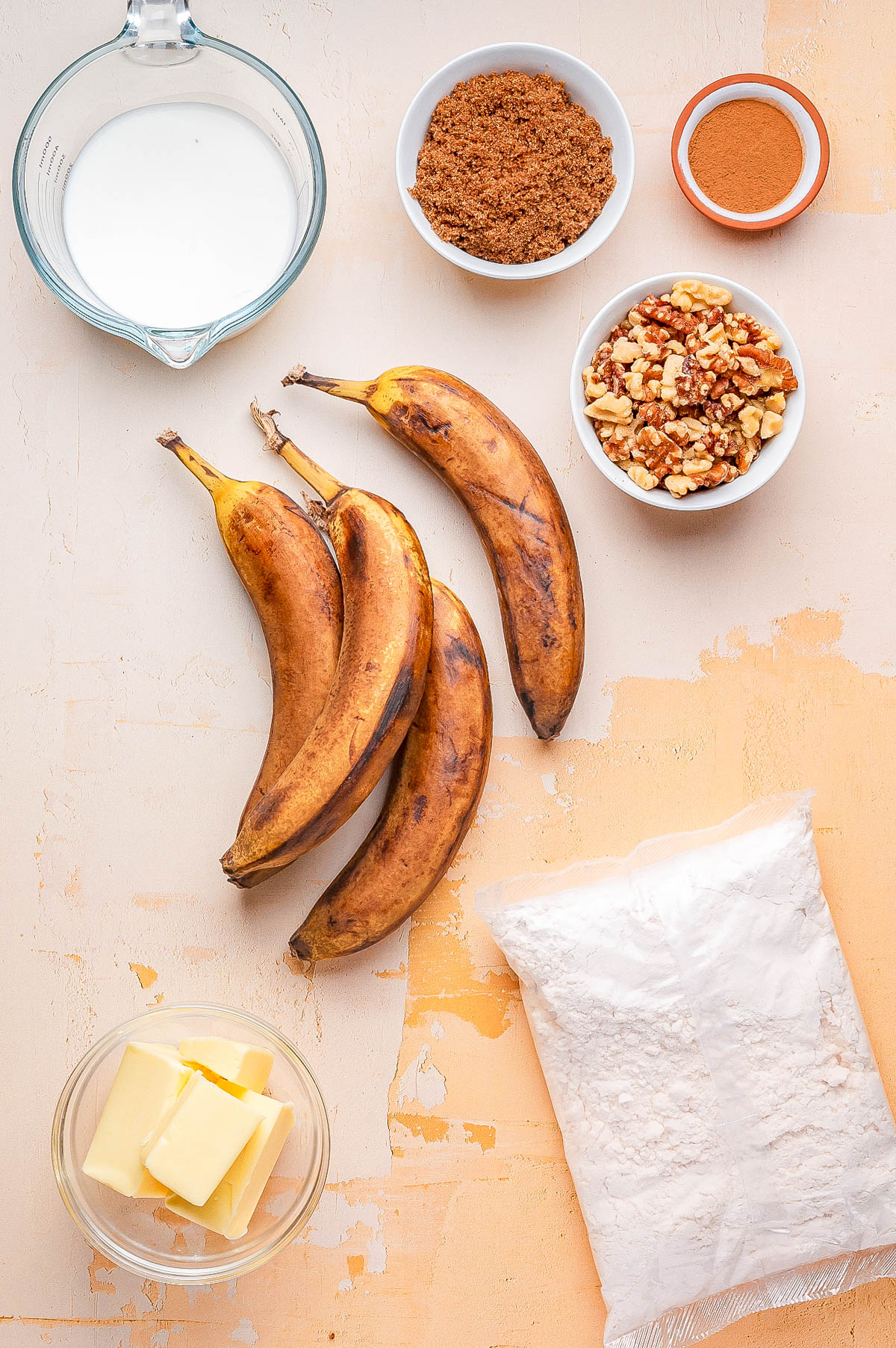 Overripe bananas, a glass measuring cup with milk, a bowl of brown sugar, cinnamon, chopped walnuts, flour in a plastic bag, and a bowl of butter arranged on a countertop.