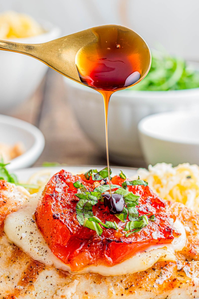 A spoon drizzling dark balsamic glaze over a piece of chicken topped with melted cheese, roasted tomato, and chopped fresh basil.