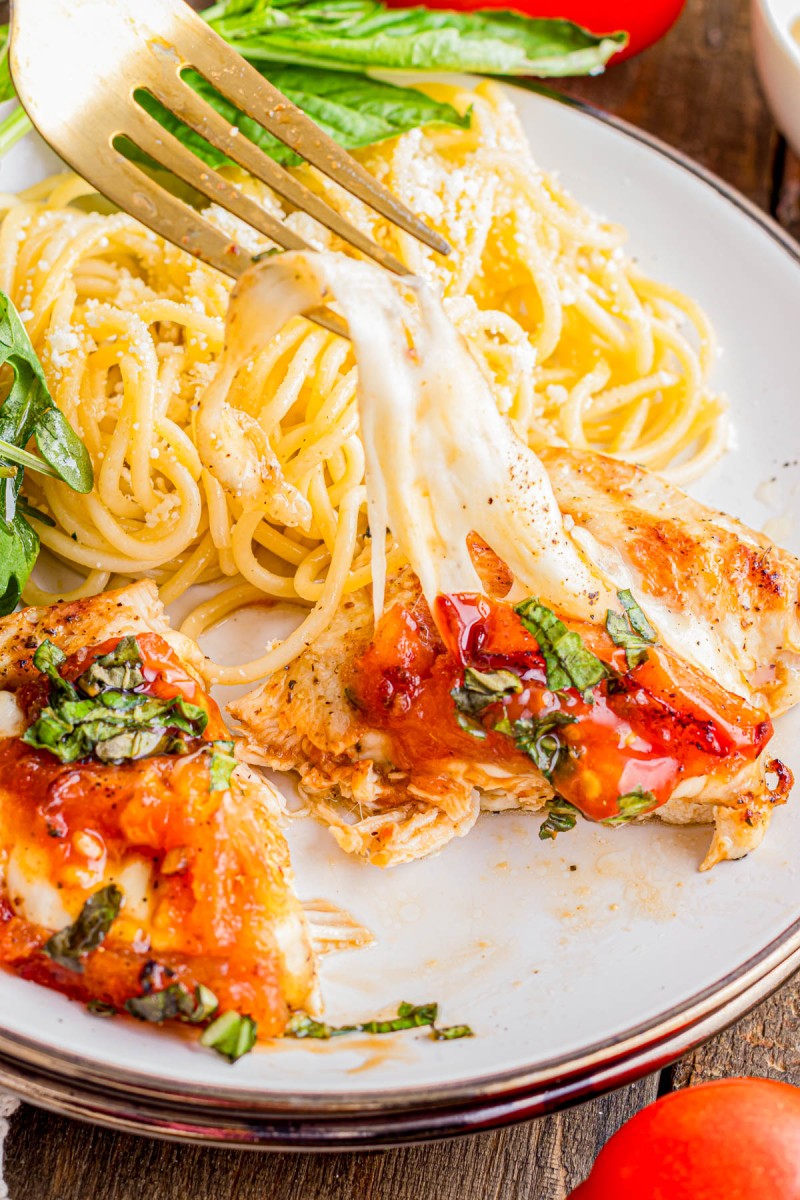 A fork lifts a cheesy piece of chicken topped with tomato sauce and basil, served with a side of spaghetti and green vegetables on a white plate.