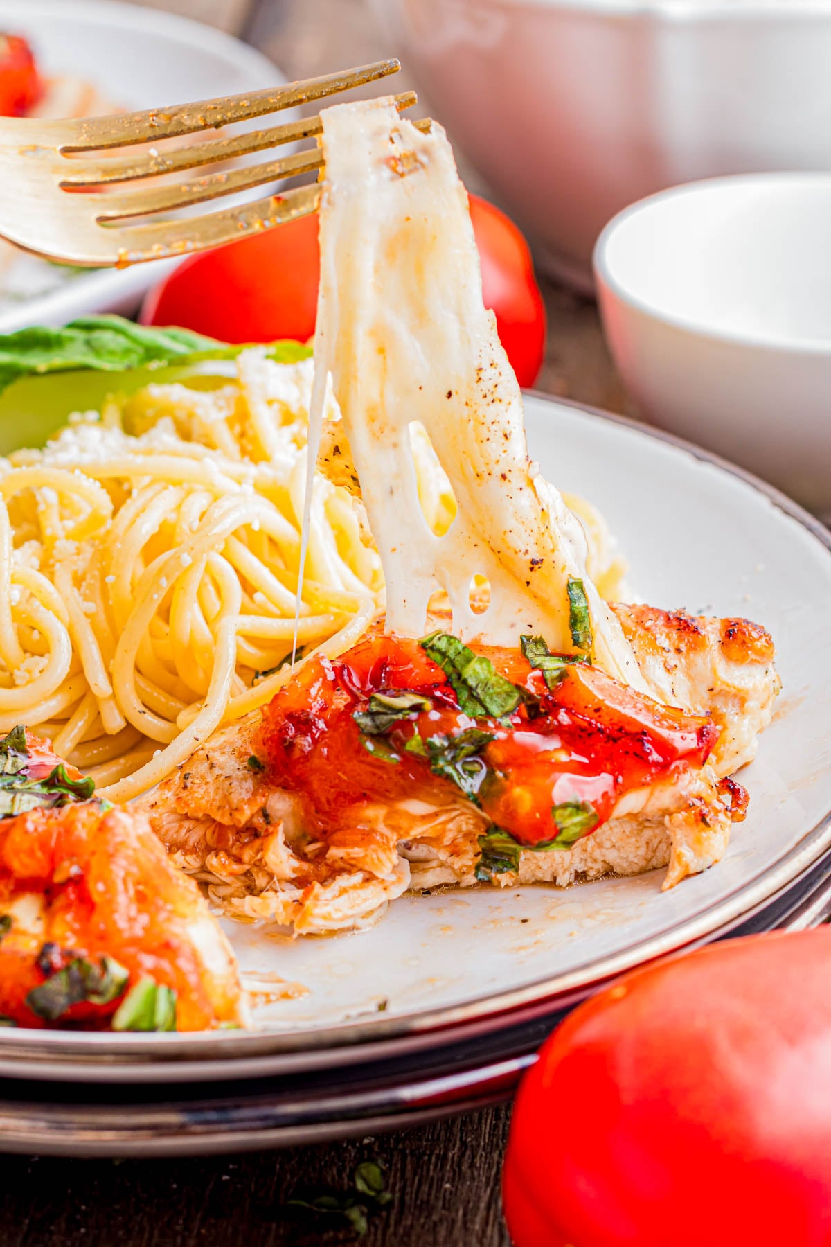 A fork lifts a piece of melted cheese from a chicken breast topped with tomatoes and herbs, served with a side of spaghetti on a white plate.