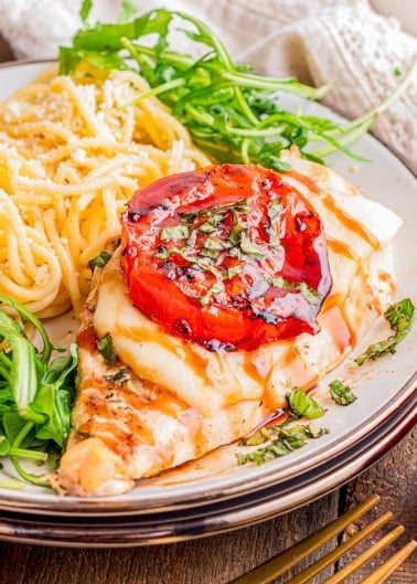 Grilled chicken breast topped with melted cheese, a roasted tomato, and herbs, served with spaghetti and arugula on a plate.