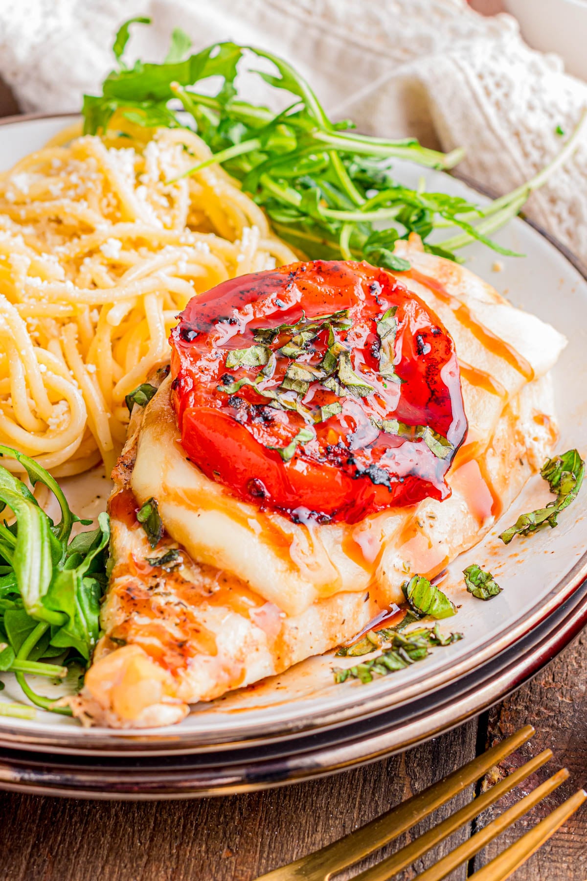 Grilled chicken breast topped with melted cheese, a roasted tomato, and herbs, served with spaghetti and arugula on a plate.