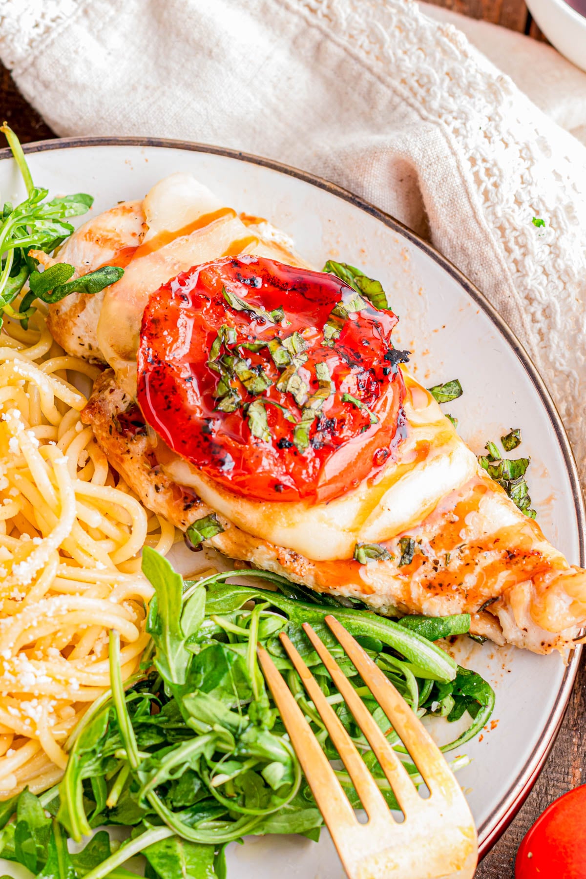 Grilled chicken breast topped with mozzarella, tomato, and herbs, served with spaghetti and a side of fresh greens on a white plate.