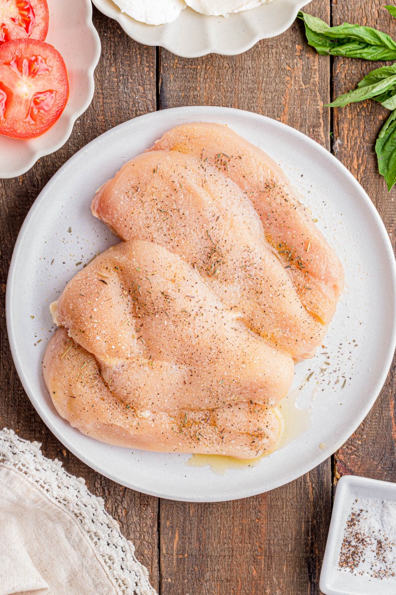 Three raw, seasoned chicken breasts on a white plate, surrounded by sliced tomatoes, fresh basil, and a container of salt on a wooden surface.