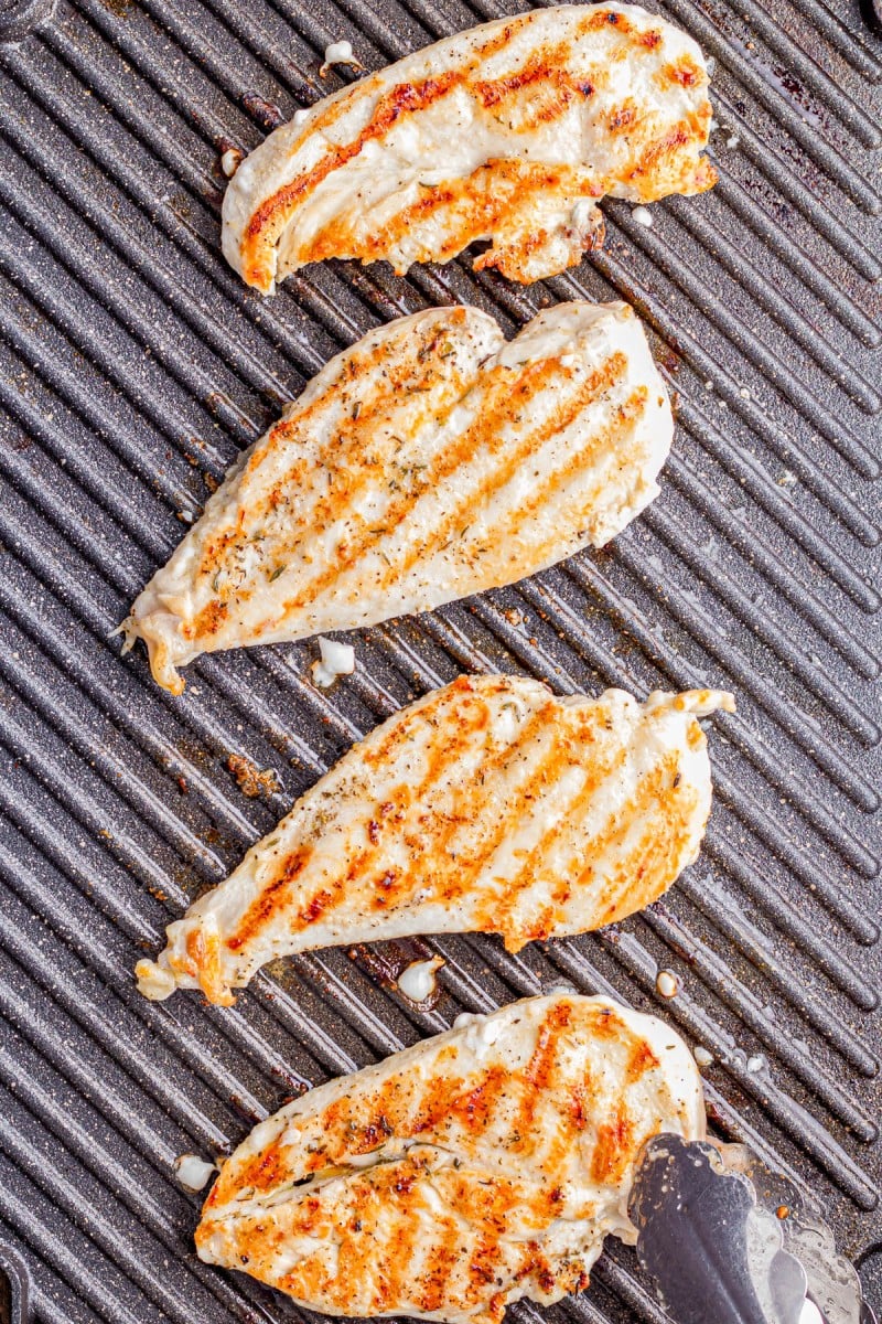 Four grilled chicken breasts with visible grill marks are cooking on a ridged grill pan, with metal tongs holding one piece.