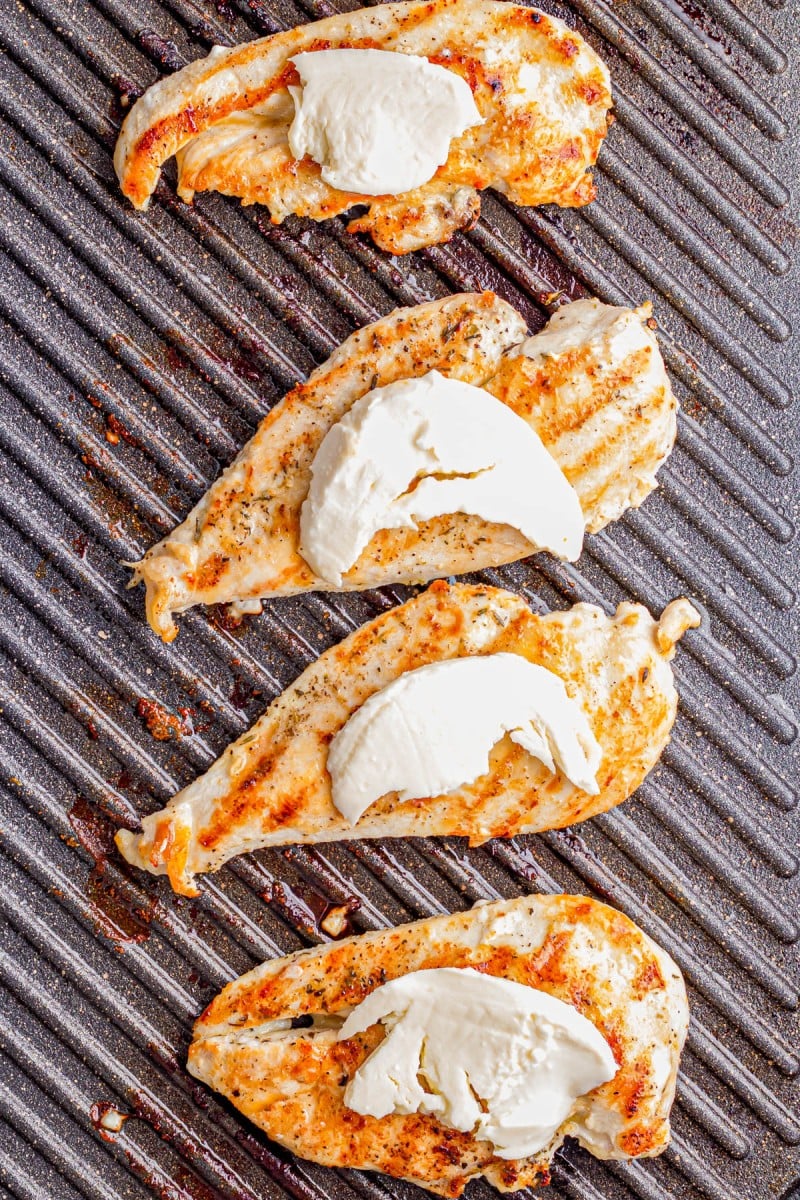 Four grilled chicken breasts topped with slices of mozzarella cheese cooking on a ribbed grill pan.
