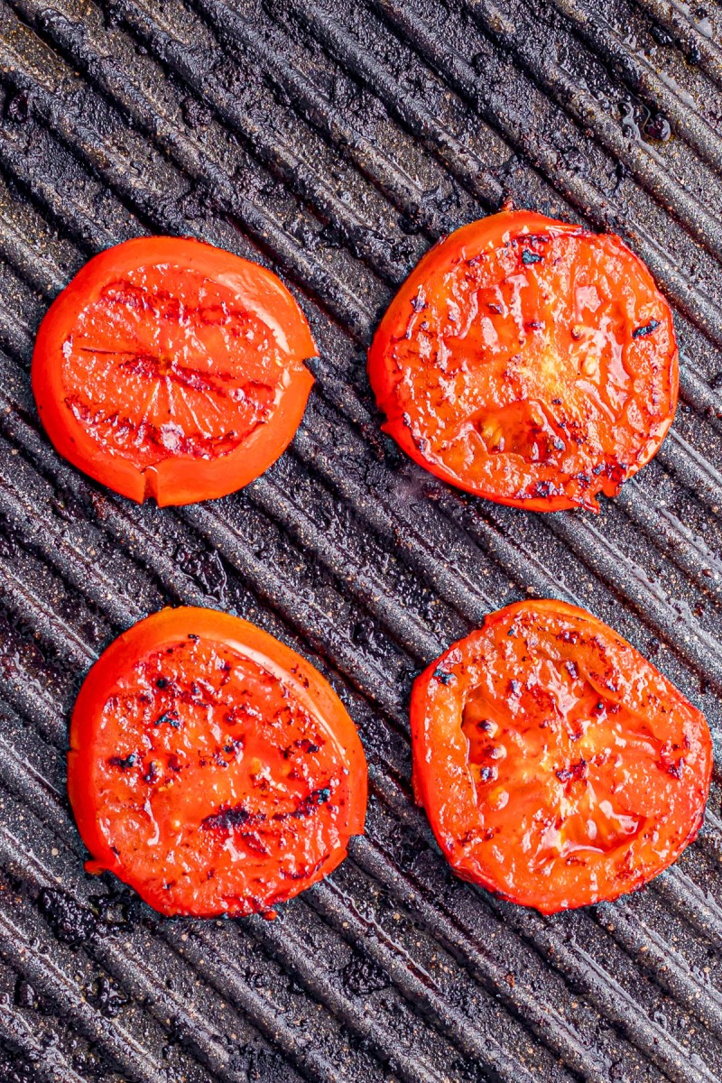 Four slices of tomato being grilled on a ridged grill surface, showing char marks and slight caramelization.