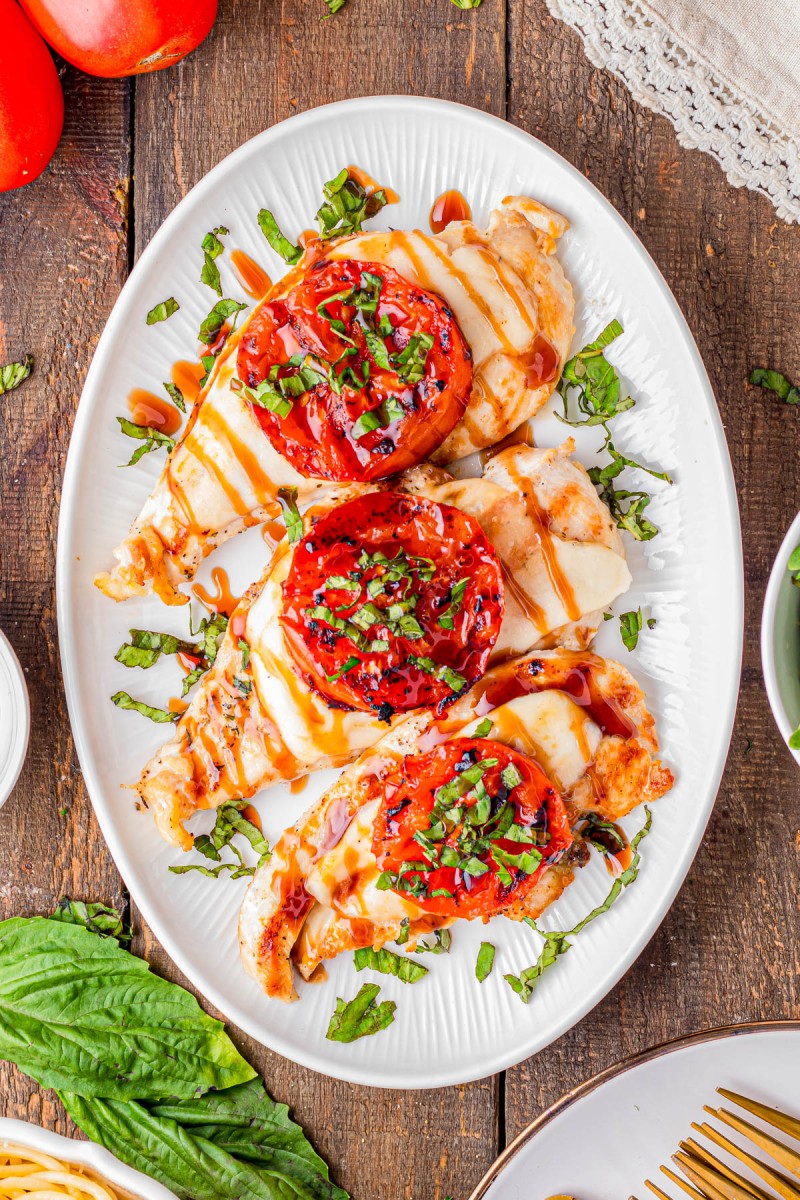 Grilled chicken breasts topped with roasted tomato slices, fresh basil, and drizzled with a balsamic glaze, served on a white oval plate.