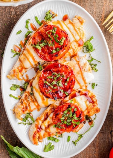Grilled chicken breasts topped with melted cheese, sliced tomatoes, chopped basil, and a drizzle of balsamic glaze on a white oval plate.