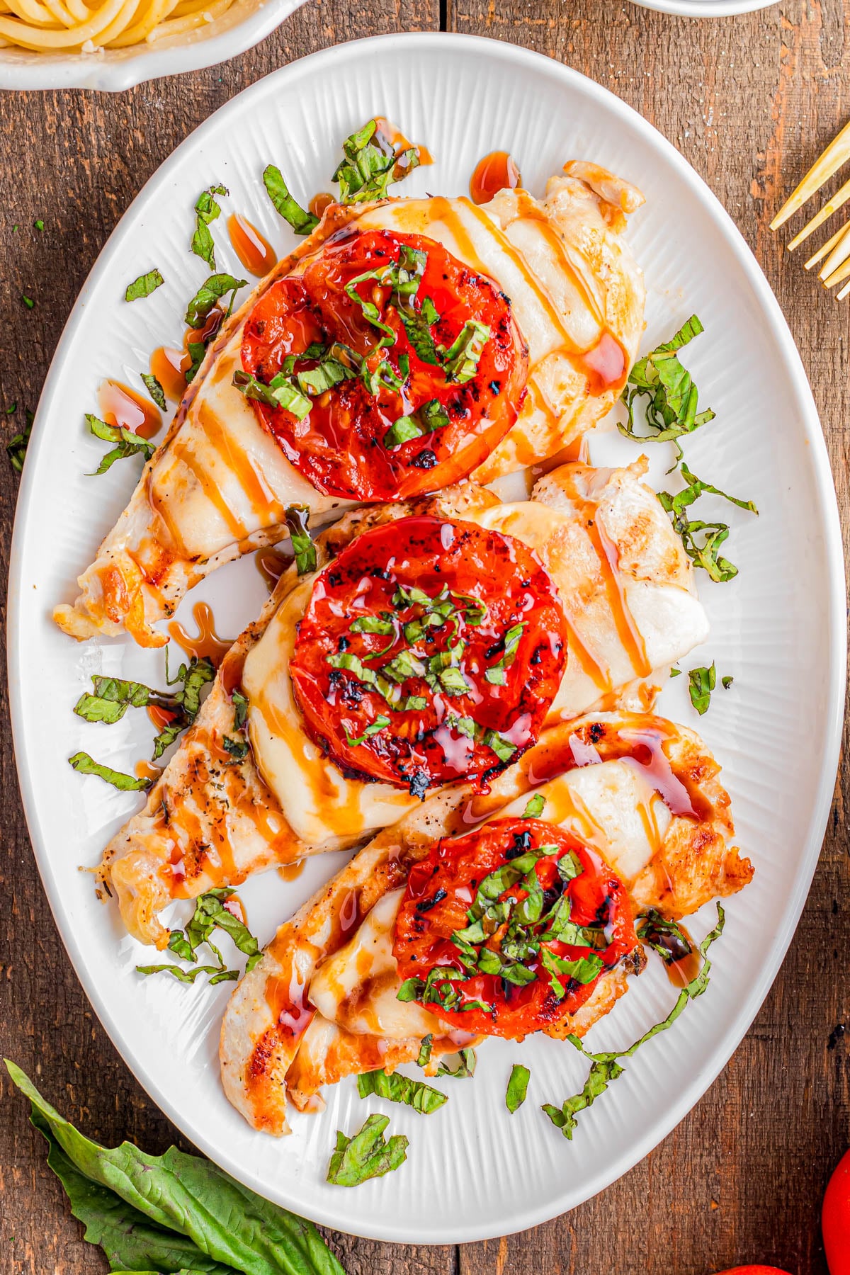 Grilled chicken breasts topped with melted cheese, sliced tomatoes, chopped basil, and a drizzle of balsamic glaze on a white oval plate.