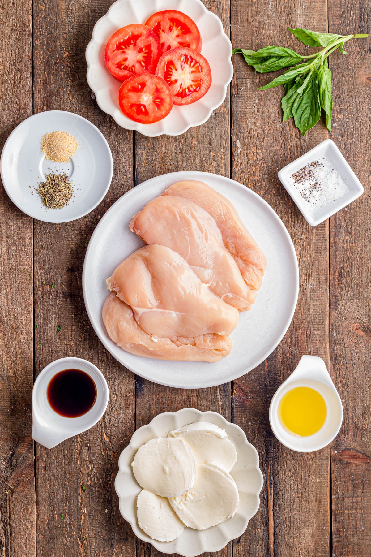 Raw chicken breasts on a plate surrounded by sliced tomatoes, mozzarella, olive oil, soy sauce, basil, and small dishes of spices on a wooden surface.