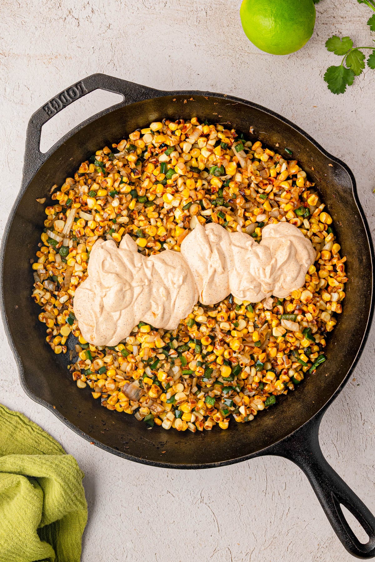 Cast iron skillet with sautรฉed corn, onions, and herbs, topped with a thick layer of light brown creamy sauce. A lime and a green towel are nearby.