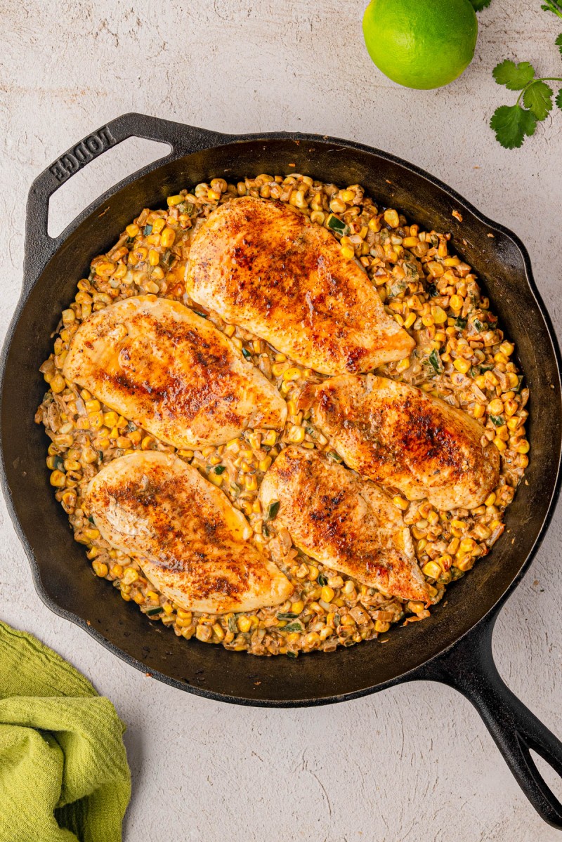 Cast iron skillet with five seasoned chicken breasts on a bed of creamy corn mixture; a lime and cilantro garnish are nearby.