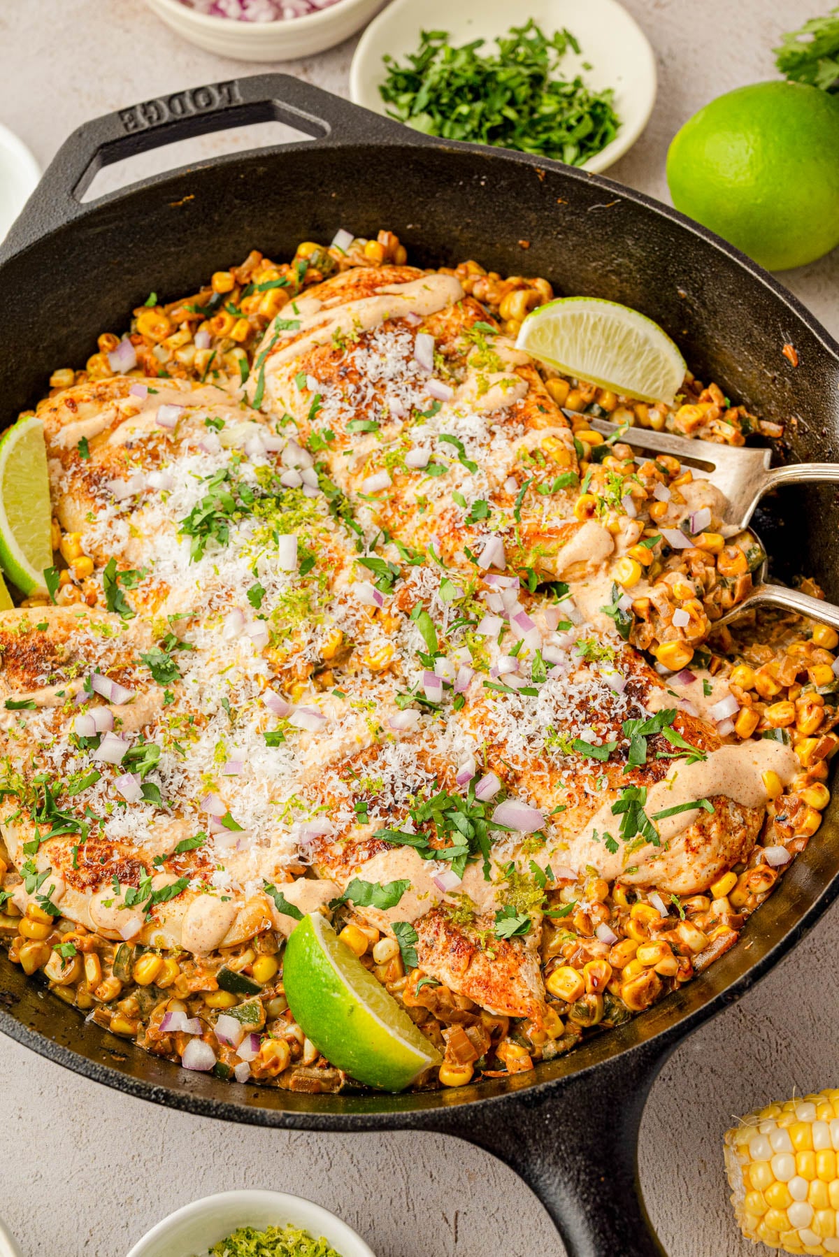 A skillet filled with seasoned chicken breasts, roasted corn, sliced red onions, grated cheese, fresh cilantro, and lime wedges.