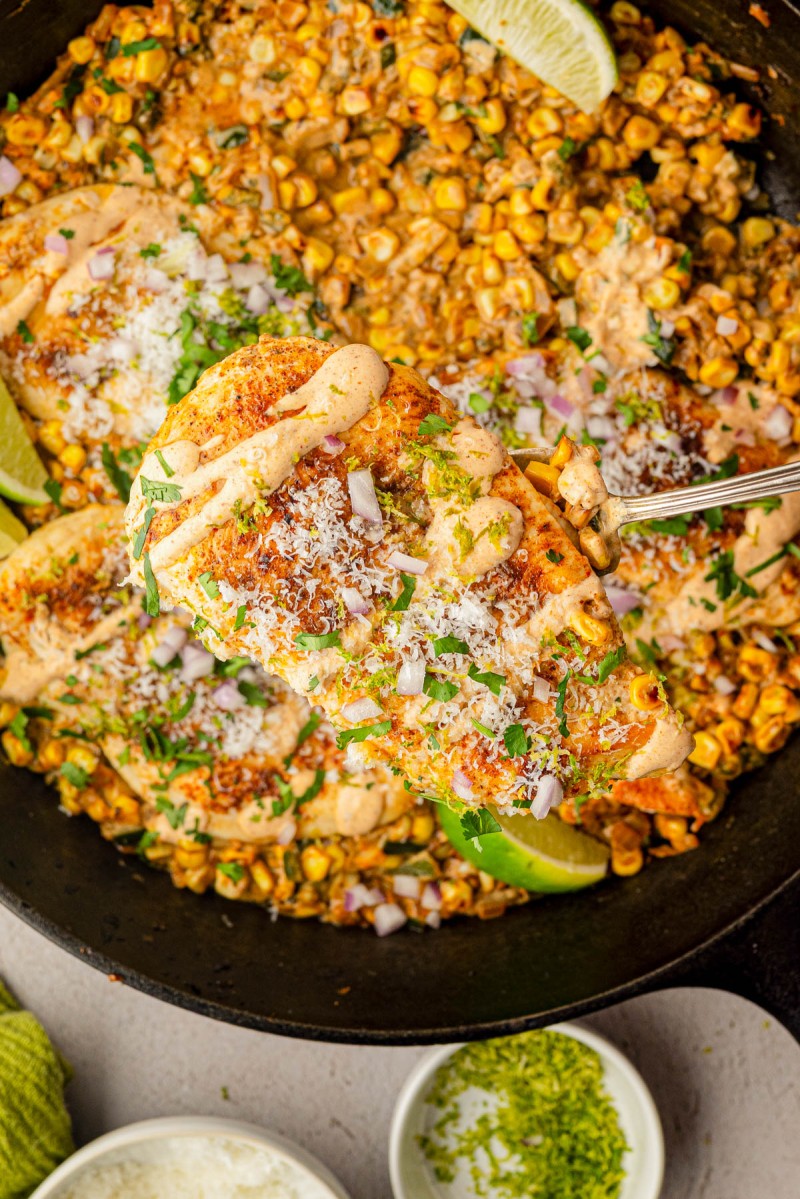 A skillet with cooked chicken breasts topped with sauce and cheese, served over corn, garnished with chopped herbs, onions, lime wedges, and grated lime zest.