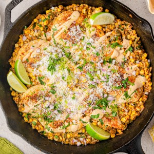 A cast iron skillet filled with grilled chicken, roasted corn, cheese, chopped herbs, lime wedges, and a creamy sauce, with small bowls of garnishes around it.