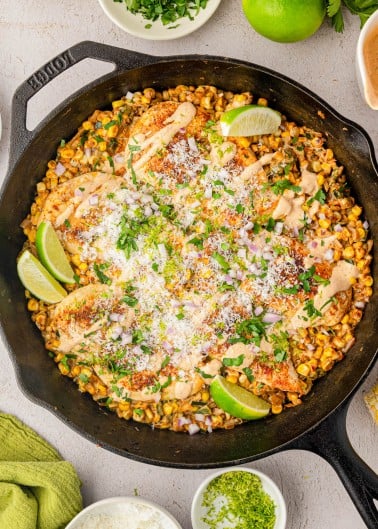 A cast iron skillet filled with grilled chicken, roasted corn, cheese, chopped herbs, lime wedges, and a creamy sauce, with small bowls of garnishes around it.