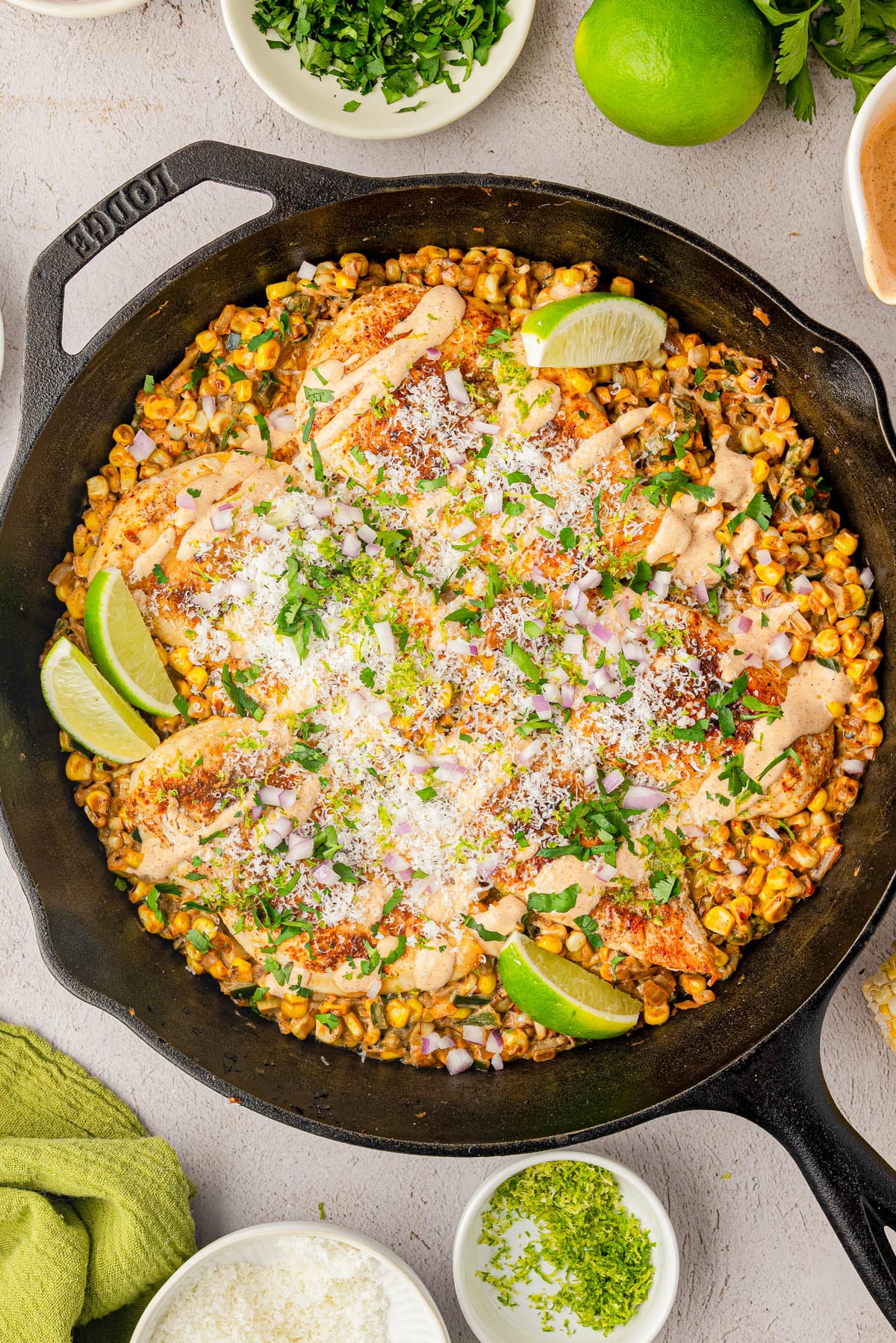 A cast iron skillet filled with grilled chicken, roasted corn, cheese, chopped herbs, lime wedges, and a creamy sauce, with small bowls of garnishes around it.