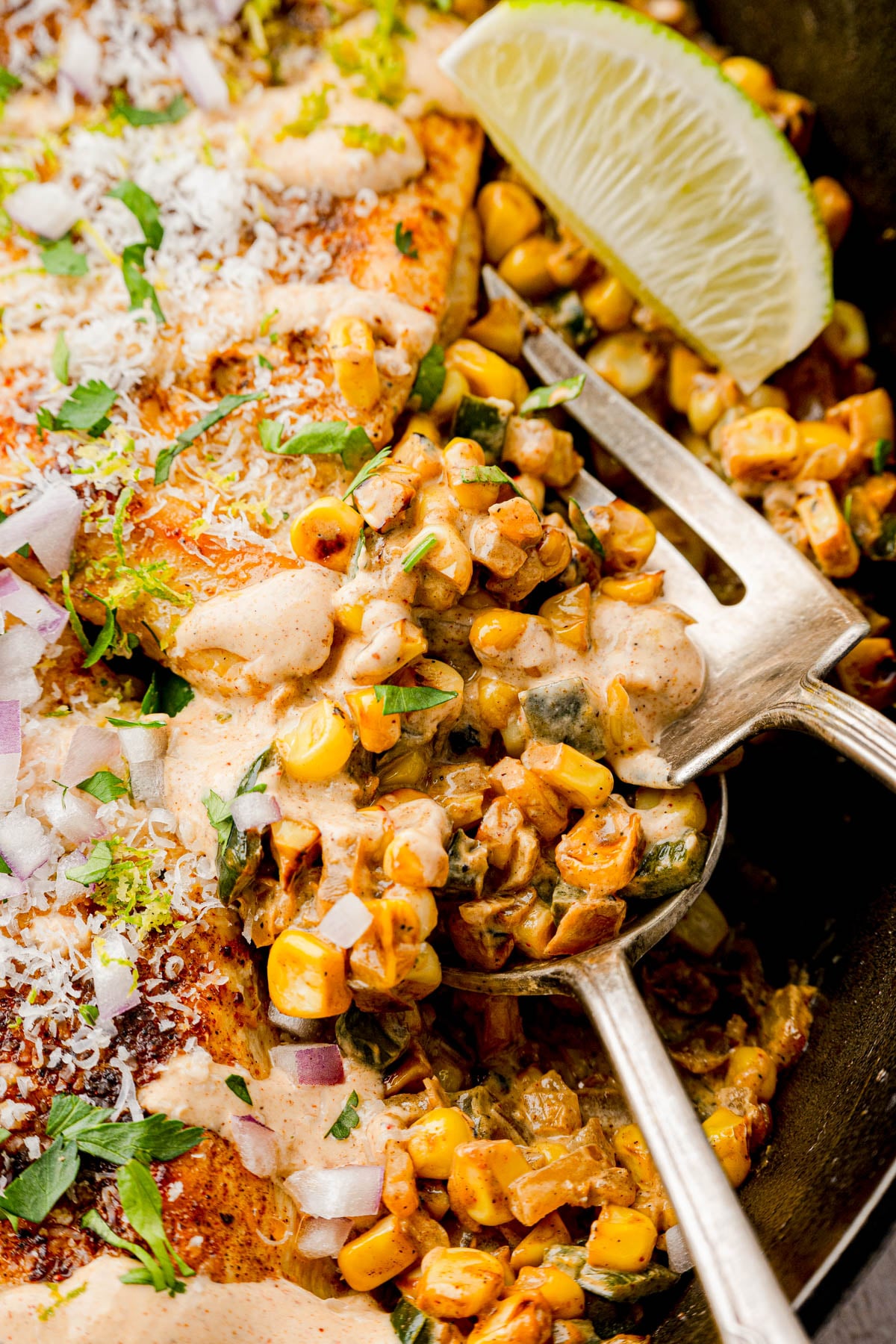 A close-up of a fork scooping creamy corn mixture with herbs and chopped onions, next to seasoned grilled chicken and a wedge of lime.
