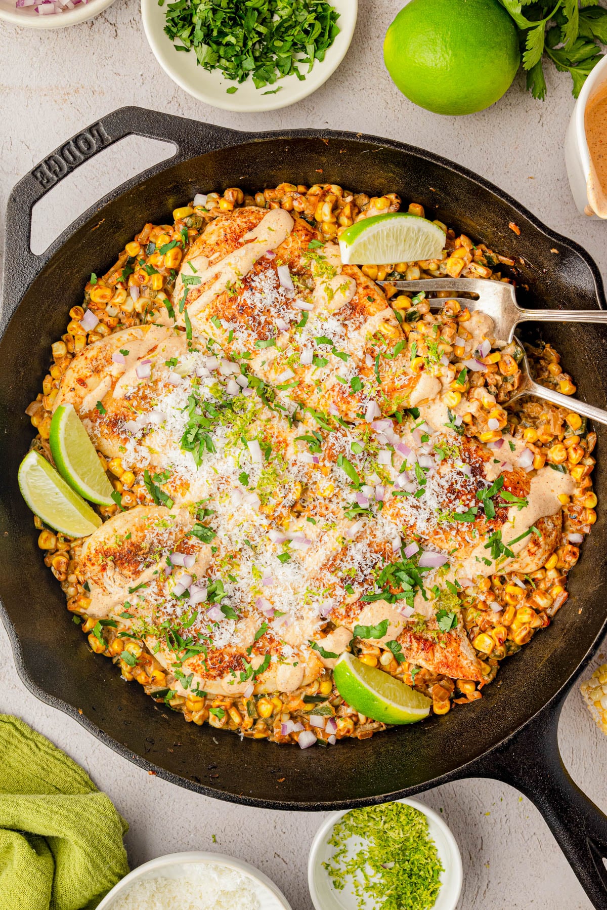 A cast iron skillet with cooked chicken breasts on a bed of seasoned corn, topped with chopped herbs, onions, grated cheese, and lime wedges, surrounded by small bowls of garnishes.