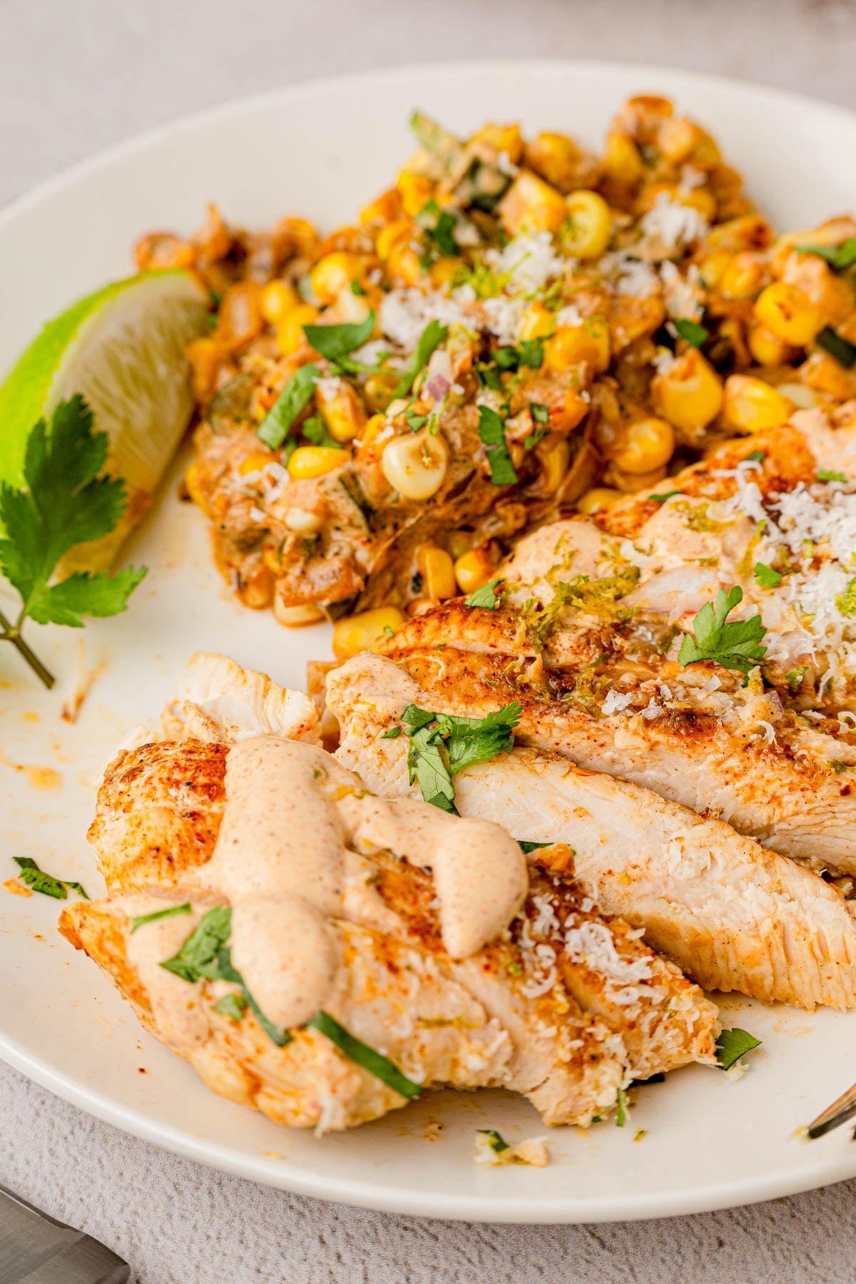Grilled chicken breast topped with creamy sauce, served with a side of seasoned corn and a lime wedge on a white plate.