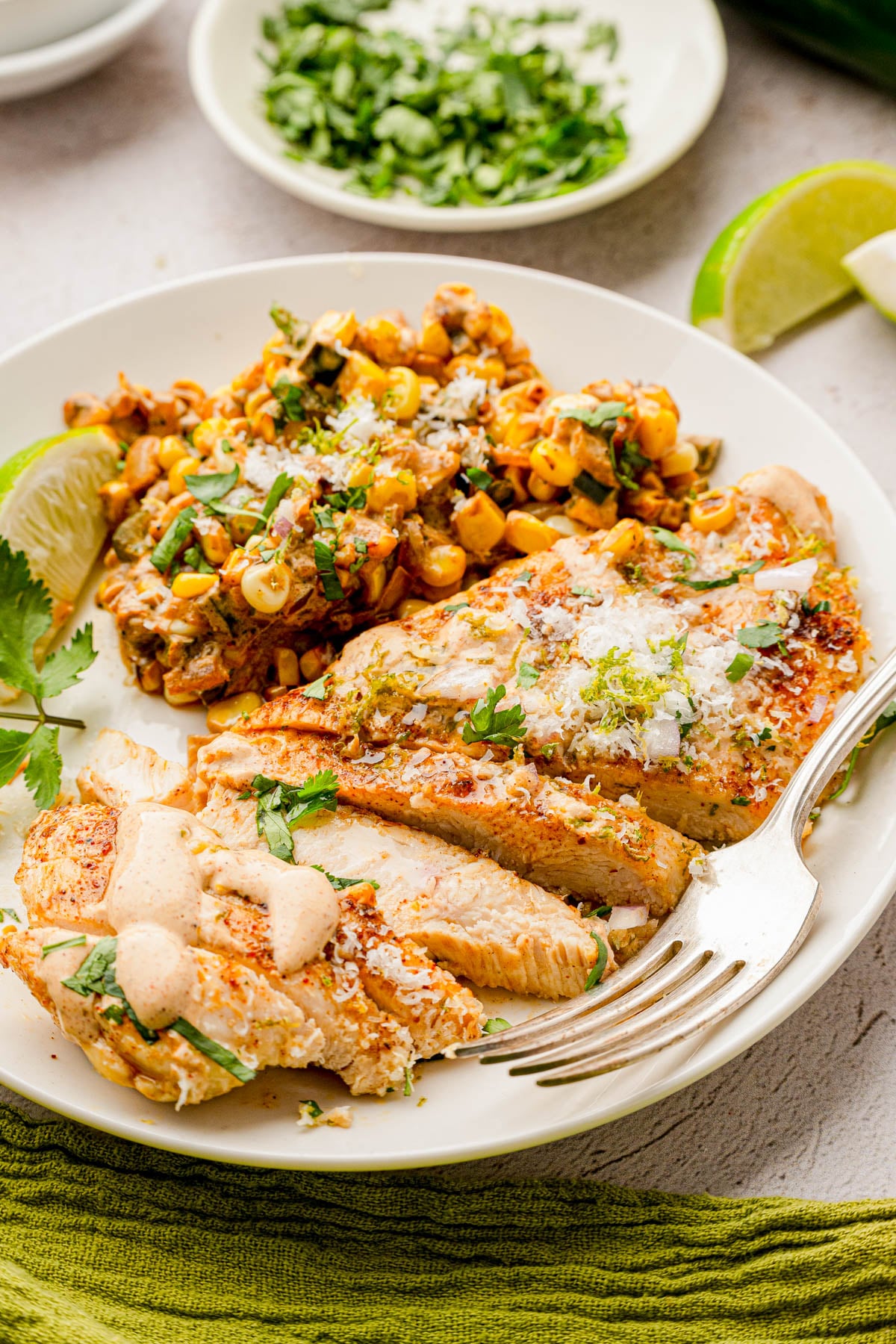 A plate with sliced grilled chicken topped with sauce, served alongside a corn and vegetable mix, garnished with herbs and a lime wedge.
