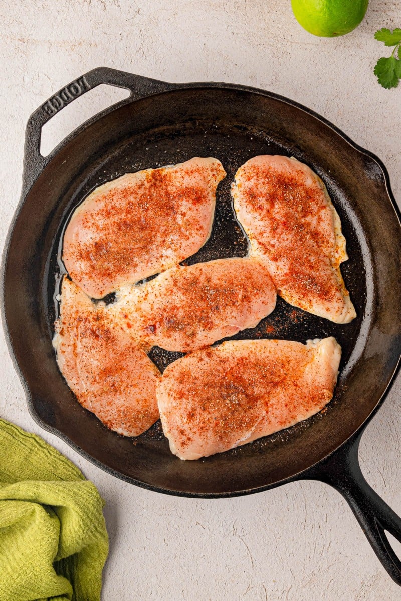 Four raw, seasoned chicken breasts in a cast iron skillet on a light surface, with a green cloth, a lime, and cilantro nearby.