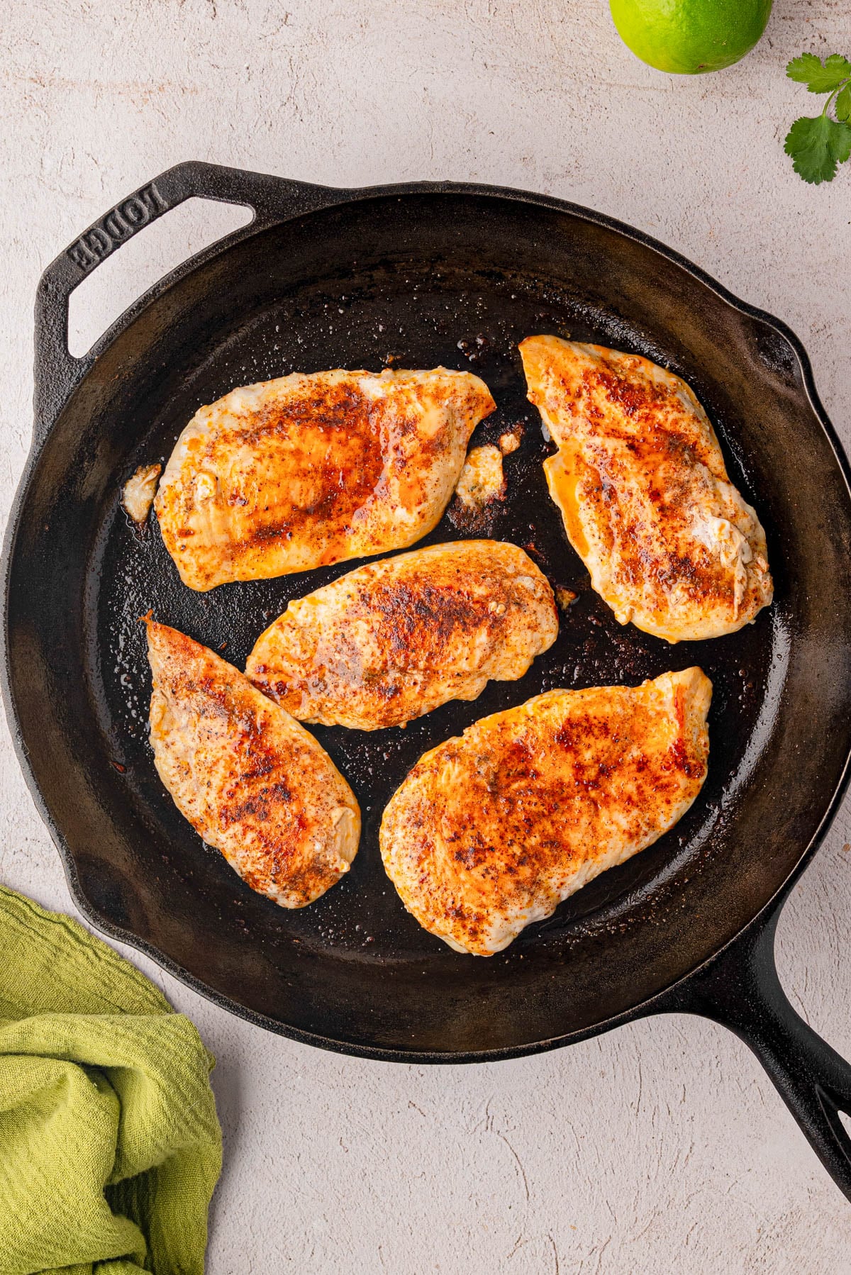 Five seasoned chicken breasts cooking in a cast iron skillet, viewed from above, with a green cloth and part of a lime visible nearby.