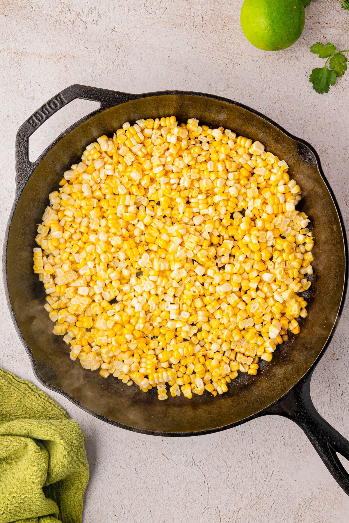 A cast iron skillet filled with cooked corn kernels sits on a light countertop, with a green lime, cilantro, and a green towel nearby.