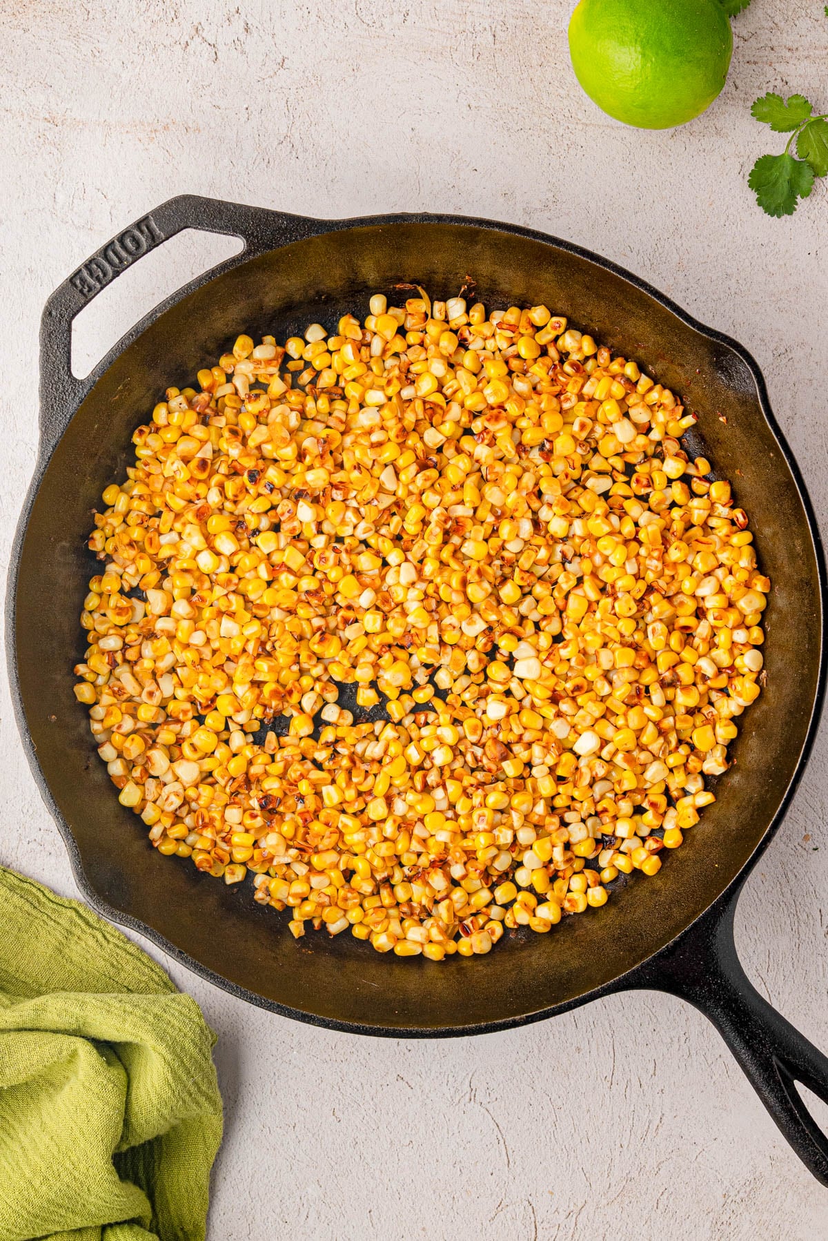 Cooked corn kernels in a cast iron skillet on a light-colored surface, with a green lime, a cilantro leaf, and a green cloth nearby.