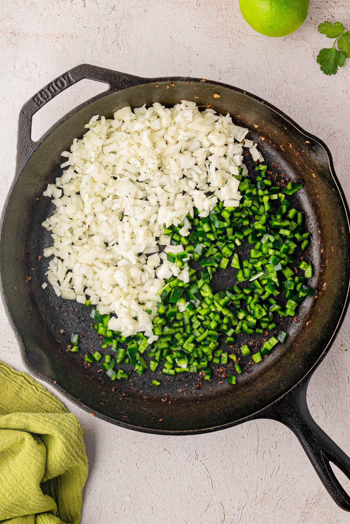 Chopped white onions and green peppers cooking in a black cast iron skillet on a light surface, with a lime, cilantro, and green towel nearby.