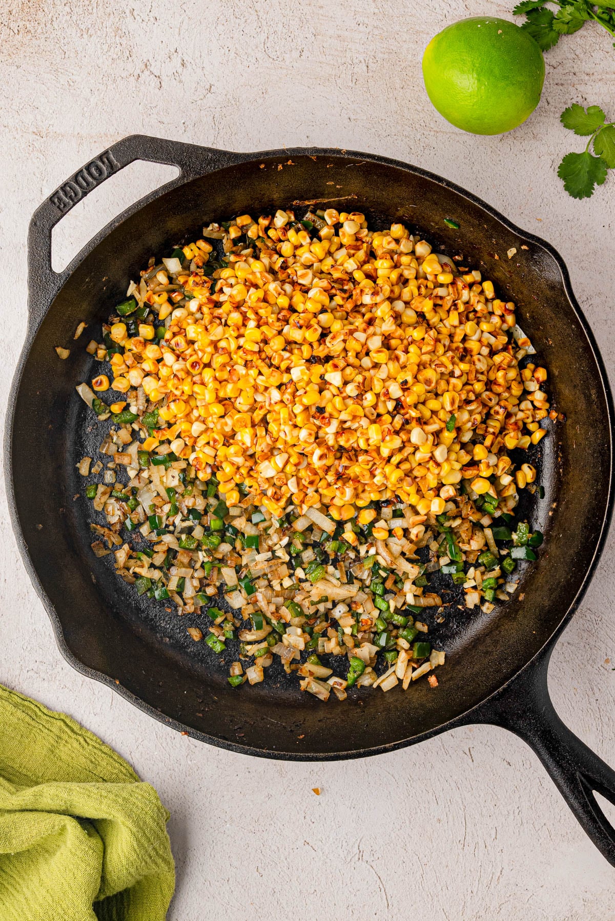 A cast iron skillet containing sautรฉed corn, onions, and chopped green peppers sits on a light surface with a lime and cilantro nearby.