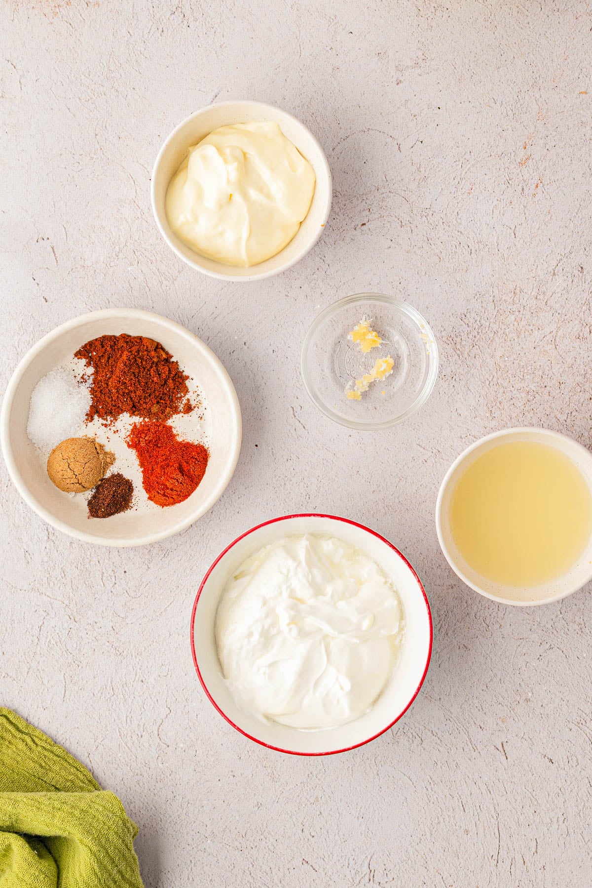 Bowls containing yogurt, mayonnaise, lemon juice, minced garlic, and a mix of spices are arranged on a light countertop with a green cloth partially visible.
