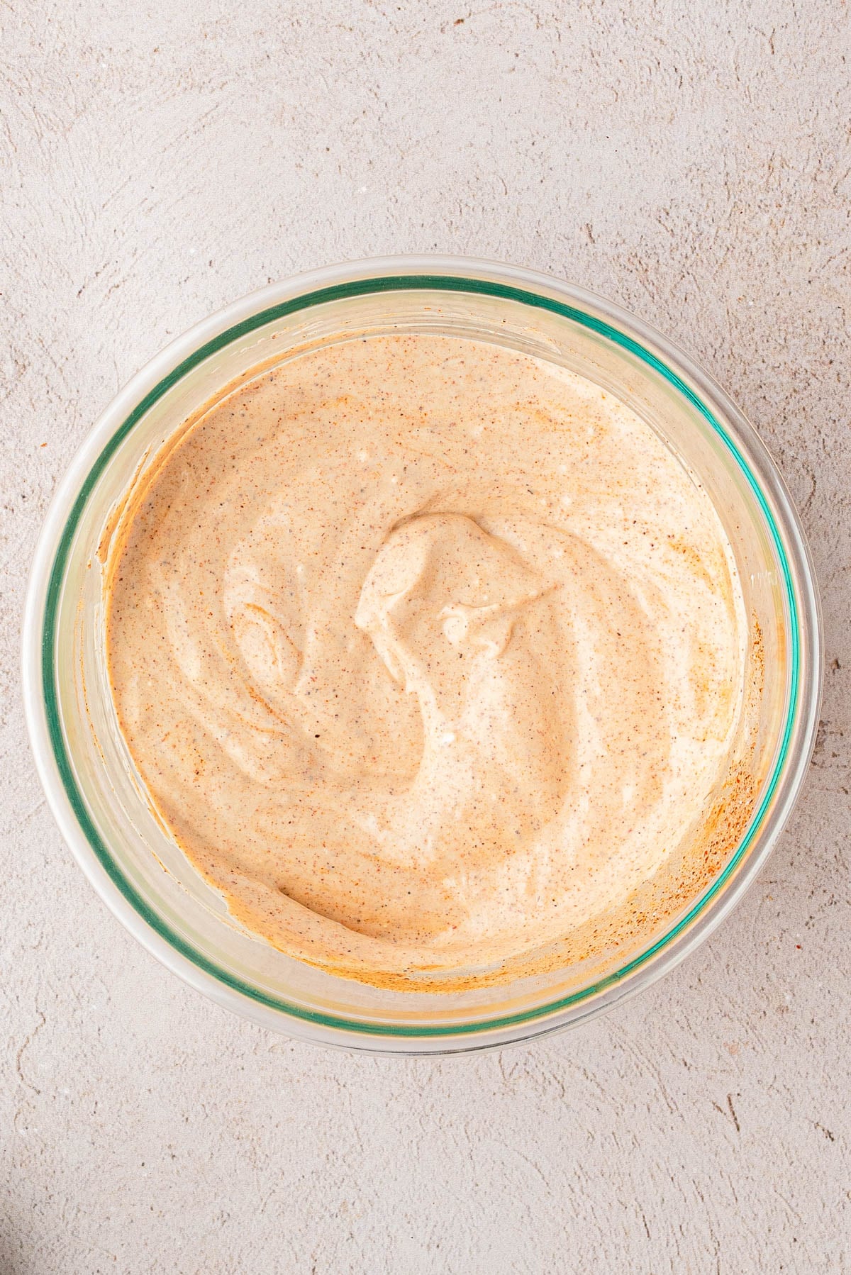 A bowl filled with a creamy, light brown sauce or dip, swirled smoothly on a textured light surface.