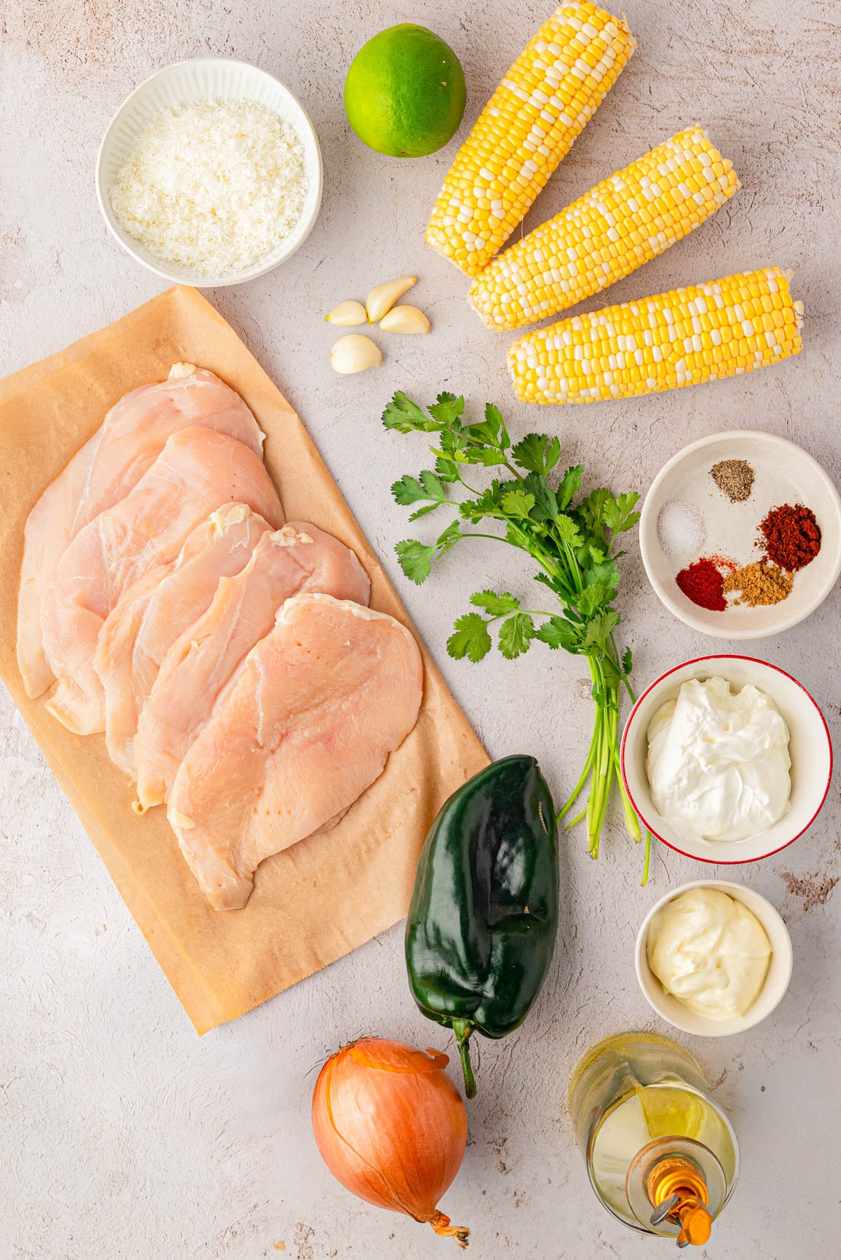 Raw chicken breasts, corn cobs, mayonnaise, crema, poblano pepper, onion, cilantro, garlic, lime, oil, grated cheese, and various spices arranged on a light surface.