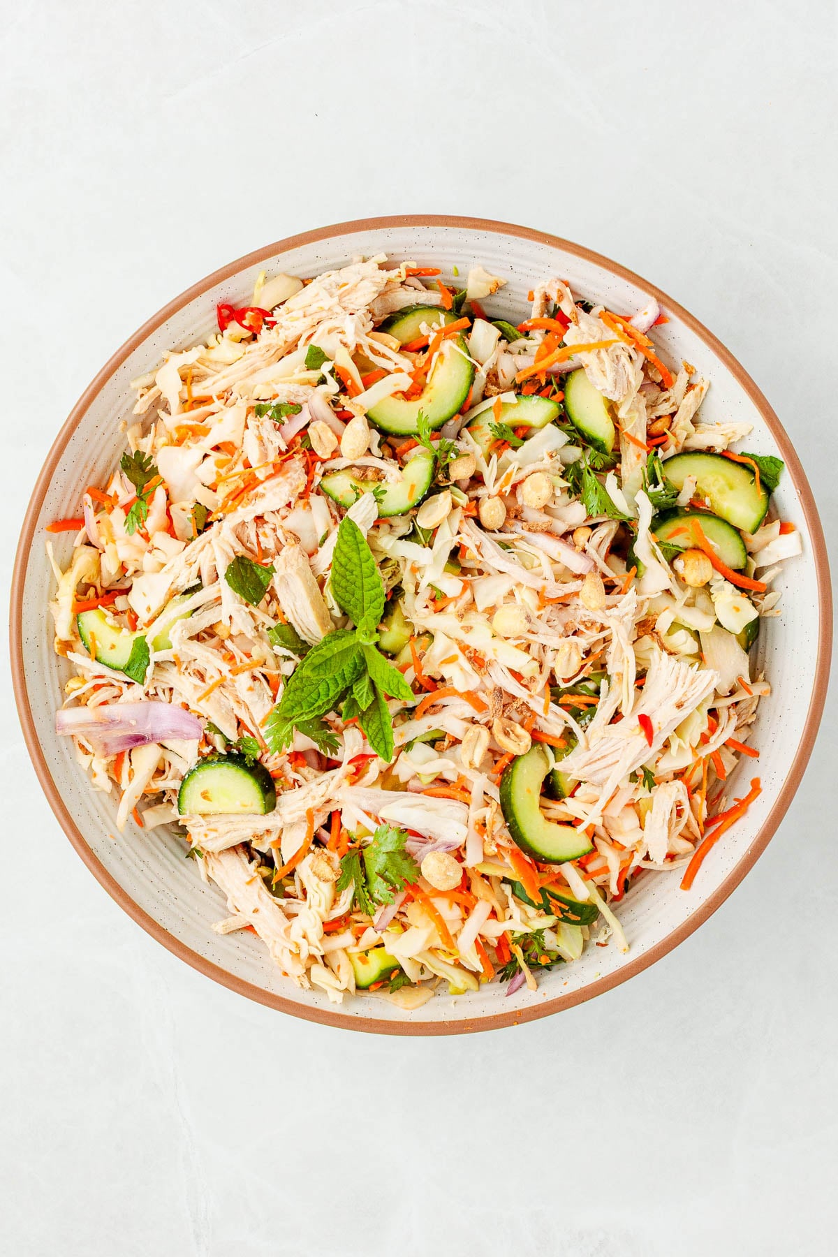 A bowl of shredded chicken salad with cucumber, carrots, red onion, chopped nuts, and fresh mint on top.