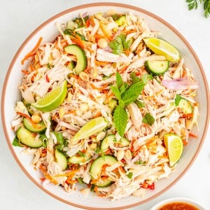 A bowl of shredded chicken salad with sliced cucumbers, carrots, red onions, peanuts, lime wedges, and fresh mint, with a bowl of dressing and serving utensils nearby.