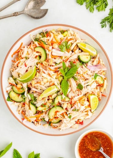 A bowl of shredded chicken salad with sliced cucumbers, carrots, red onions, peanuts, lime wedges, and fresh mint, with a bowl of dressing and serving utensils nearby.