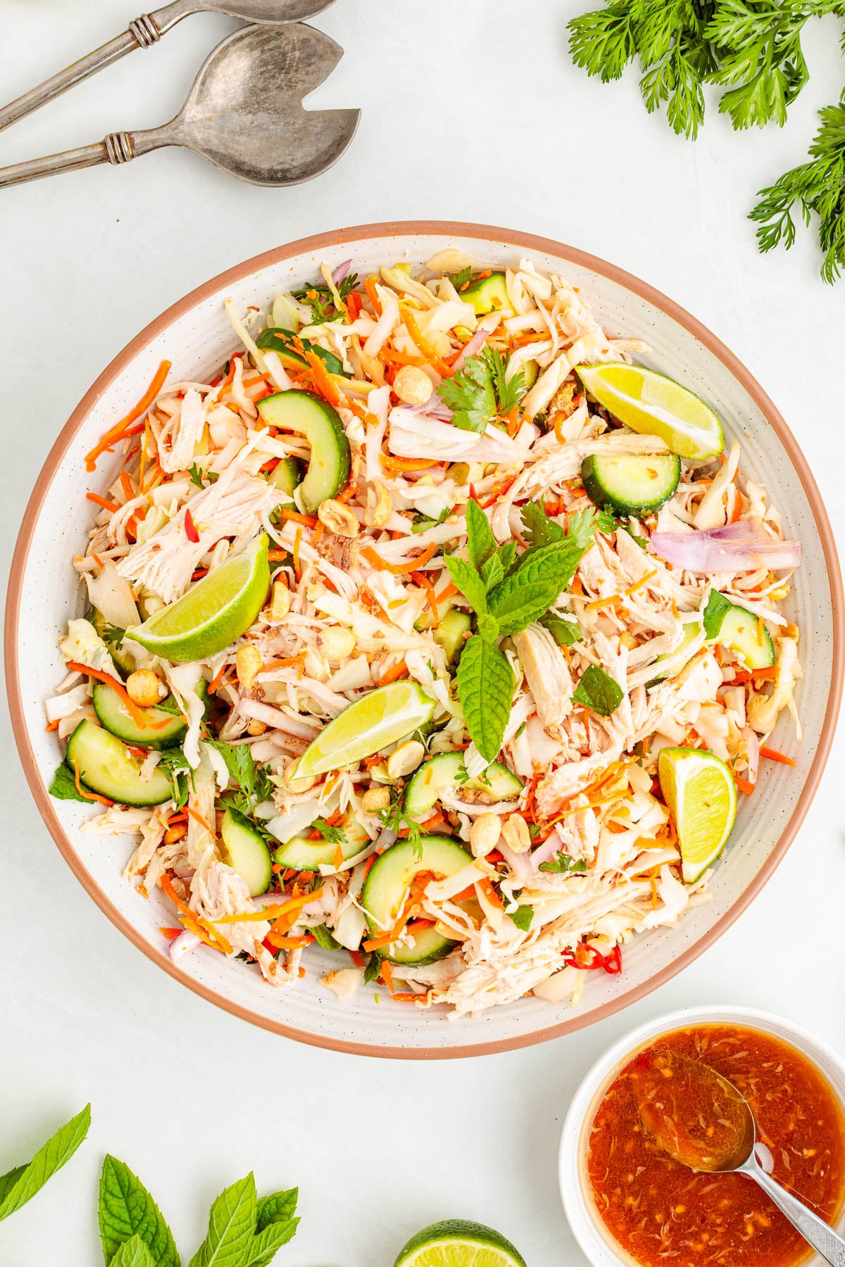 A bowl of shredded chicken salad with sliced cucumbers, carrots, red onions, peanuts, lime wedges, and fresh mint, with a bowl of dressing and serving utensils nearby.