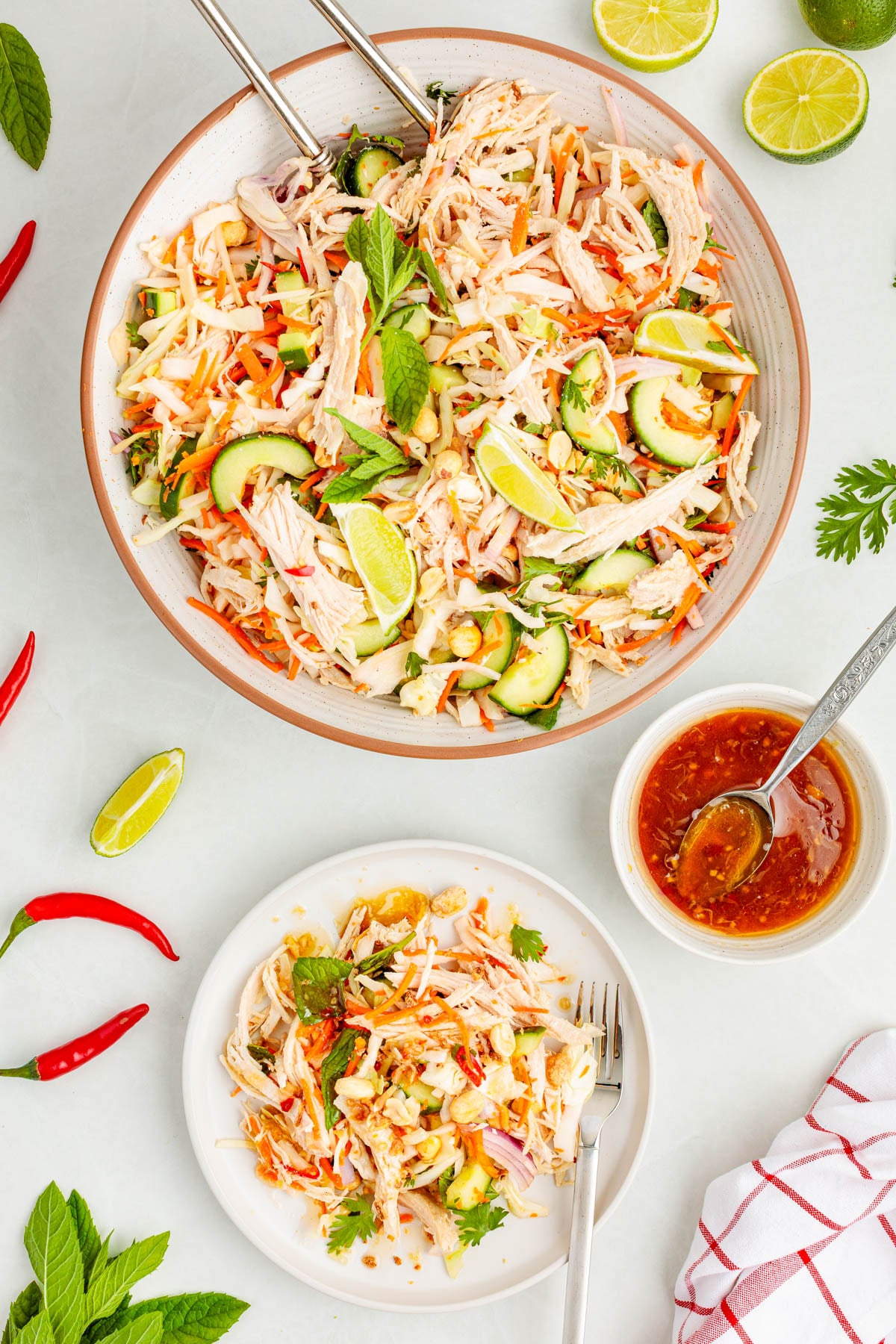 A large bowl and a plate of shredded chicken salad with cucumbers, carrots, herbs, lime wedges, and peanuts, served with a side of orange dressing.