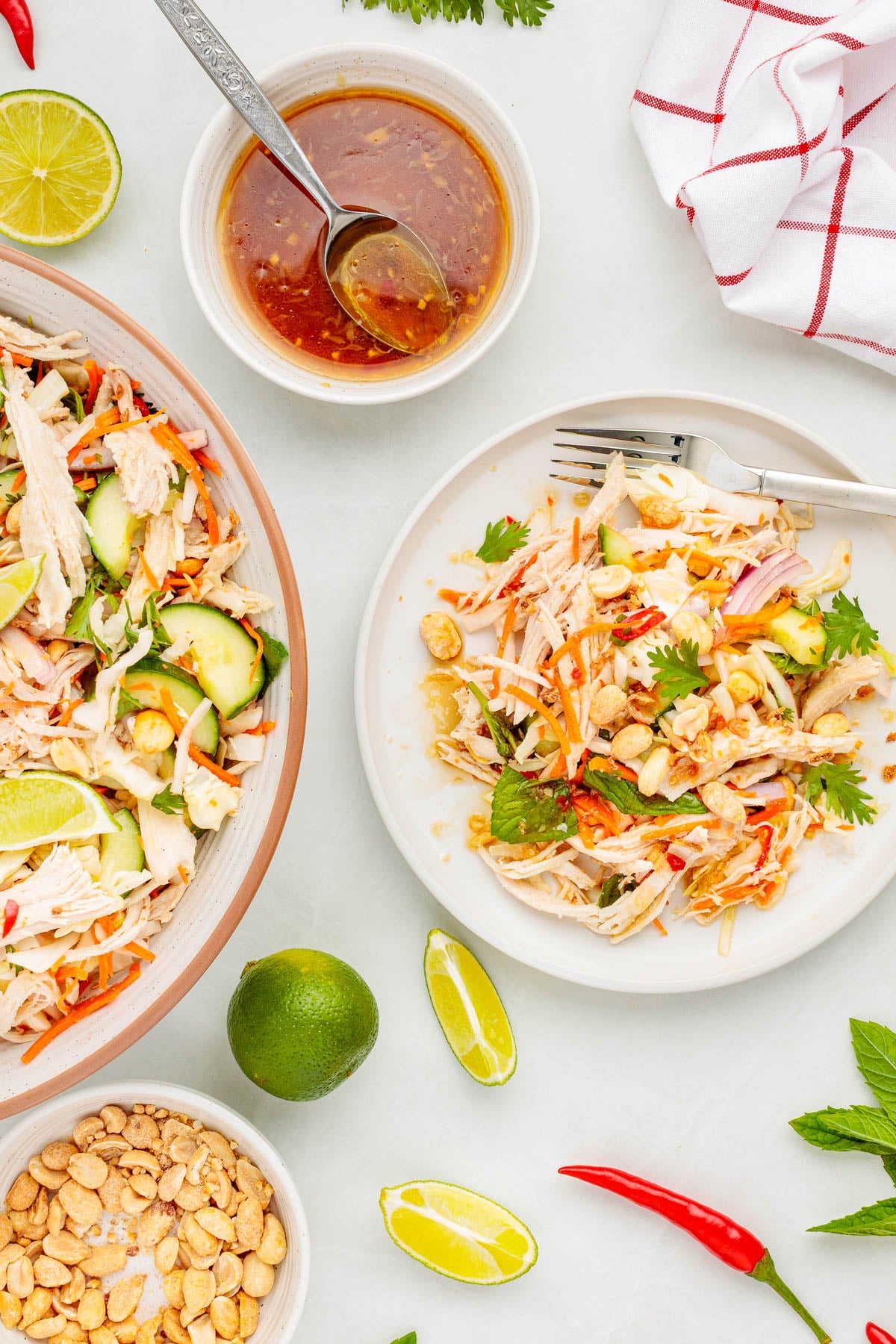 A plate of shredded chicken salad with cucumbers, carrots, peanuts, herbs, lime wedges, and a bowl of dressing. Extra peanuts, lime, and chili peppers are scattered on the table.
