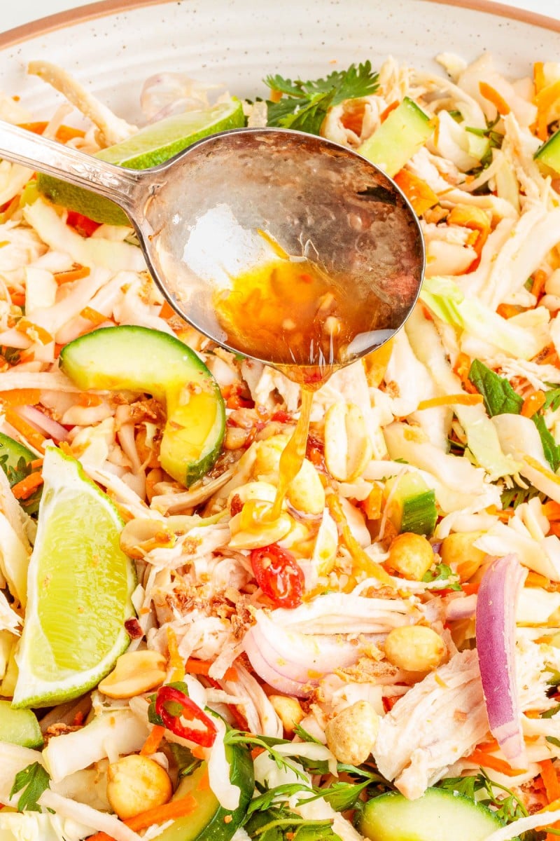 A spoon pours dressing over a salad with shredded chicken, sliced avocado, lime wedges, red onions, chilies, and greens.
