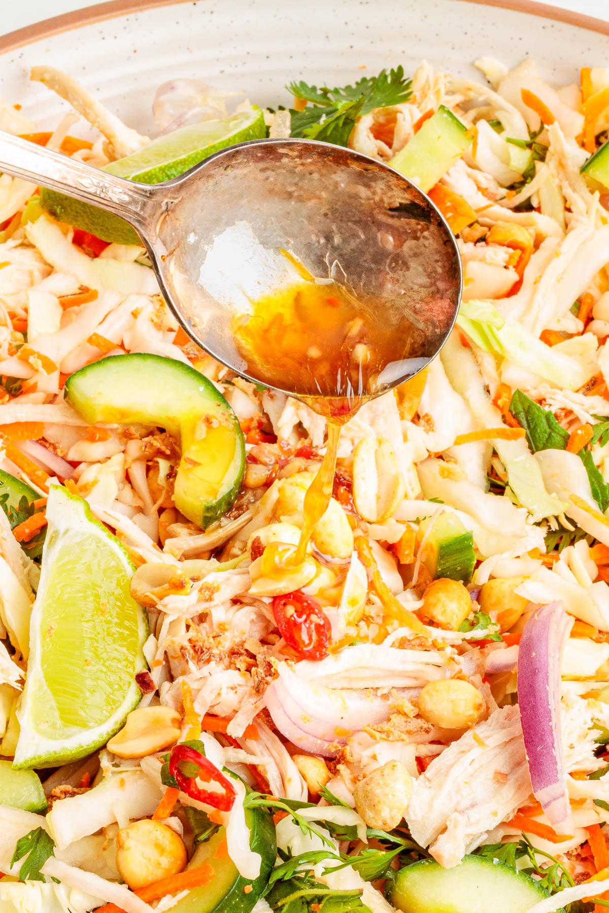 A spoon pours dressing over a salad with shredded chicken, sliced avocado, lime wedges, red onions, chilies, and greens.
