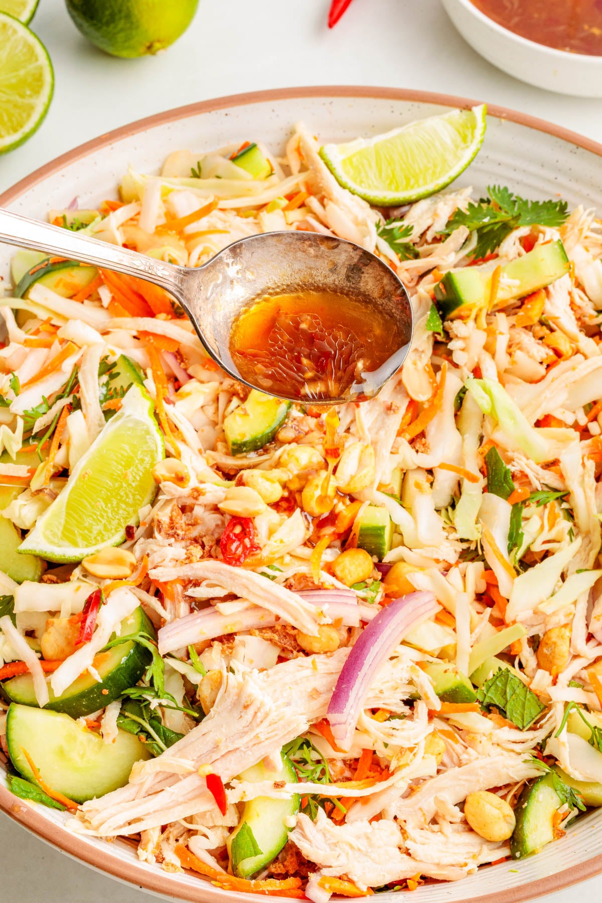 A large bowl of shredded chicken salad with sliced cucumbers, red onions, lime wedges, peanuts, and cilantro, being drizzled with dressing from a ladle.