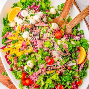 Oval platter of mixed salad with lettuce, tomatoes, mozzarella balls, sliced salami, yellow peppers, shredded cheese, and lemon wedges. Wooden salad servers rest on the dish.