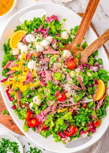 Oval platter of mixed salad with lettuce, tomatoes, mozzarella balls, sliced salami, yellow peppers, shredded cheese, and lemon wedges. Wooden salad servers rest on the dish.