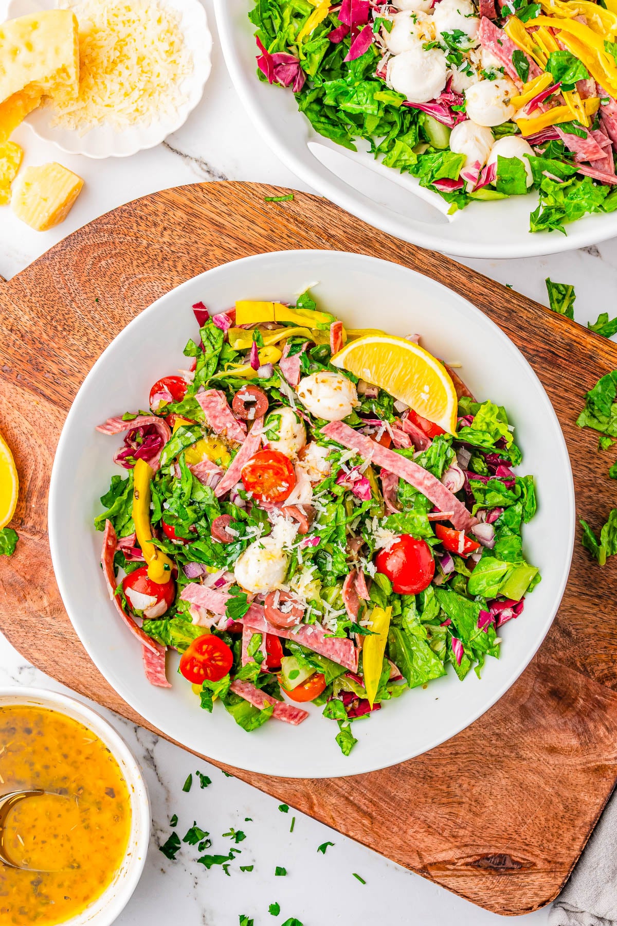 A bowl of chopped salad with lettuce, cherry tomatoes, mozzarella balls, salami strips, pepperoncini, and a lemon wedge, set on a wooden board with dressing and parmesan nearby.
