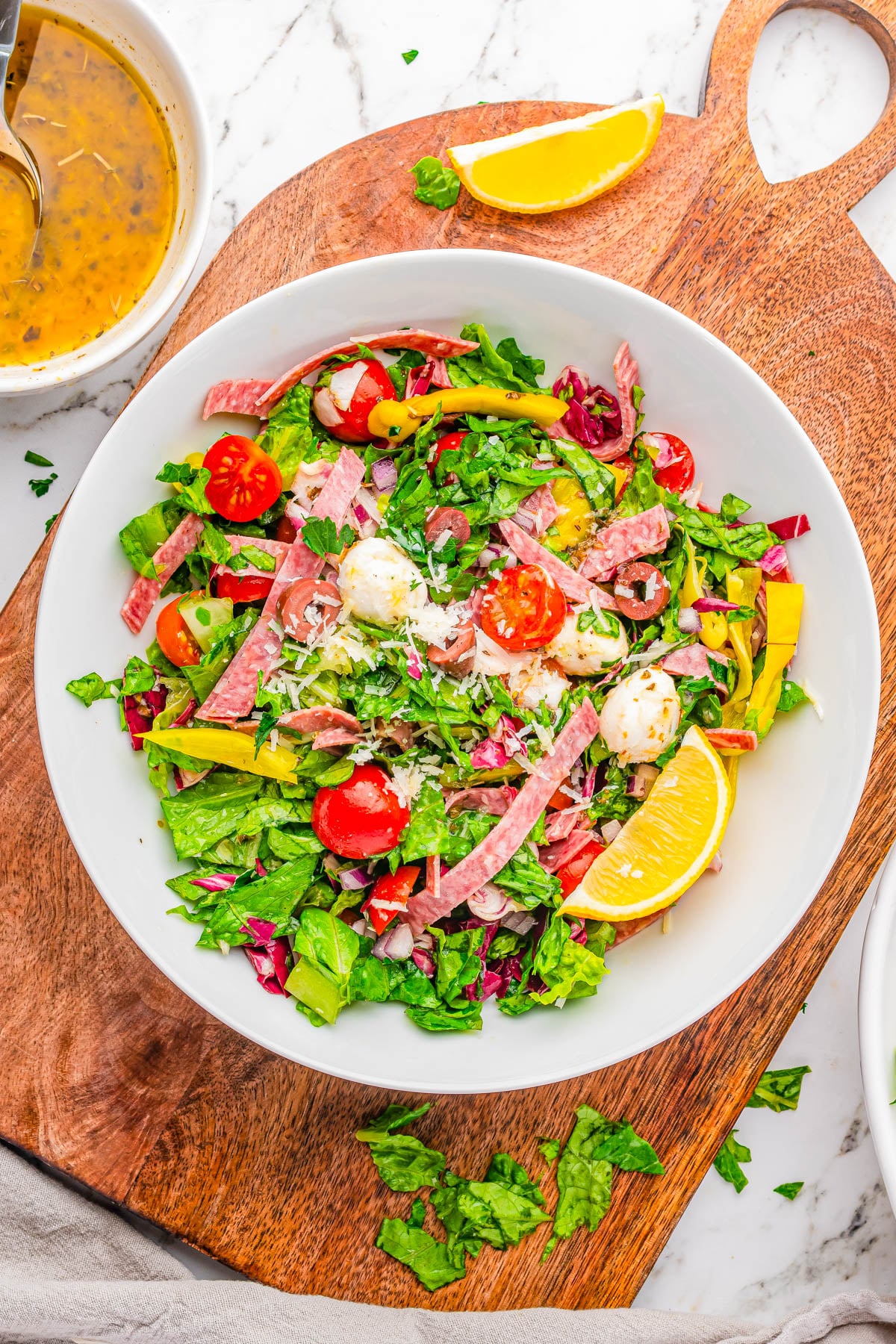 A bowl of chopped salad with lettuce, cherry tomatoes, mozzarella balls, salami strips, yellow pepper, and lemon wedge on a wooden board. Dressing in a small bowl nearby.