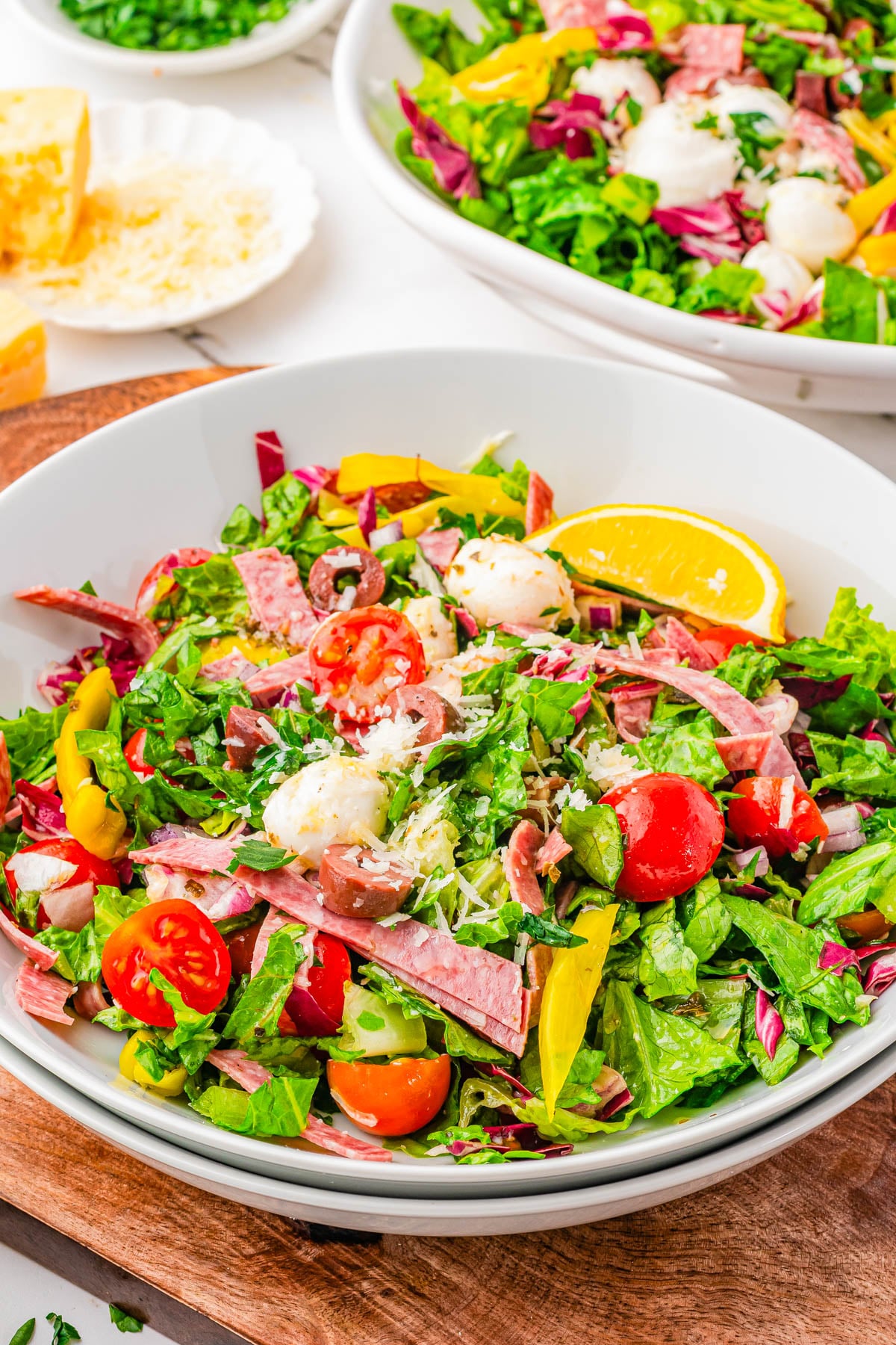 A bowl of chopped salad with lettuce, cherry tomatoes, mozzarella balls, salami slices, olives, pepperoncini, and a lemon wedge. Another salad bowl is visible in the background.